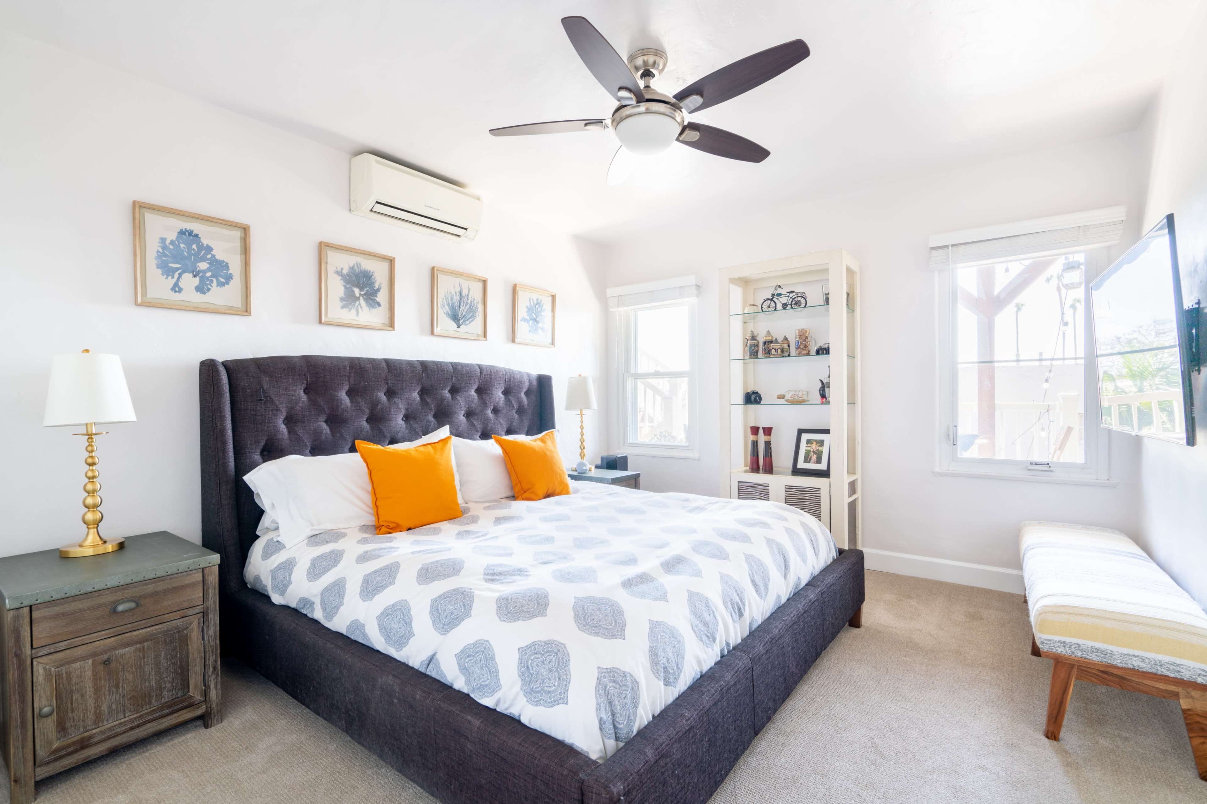 The image shows a well-lit bedroom featuring a large bed with patterned bedding, two orange pillows, a wooden nightstand, a bench, and framed artwork on the walls.