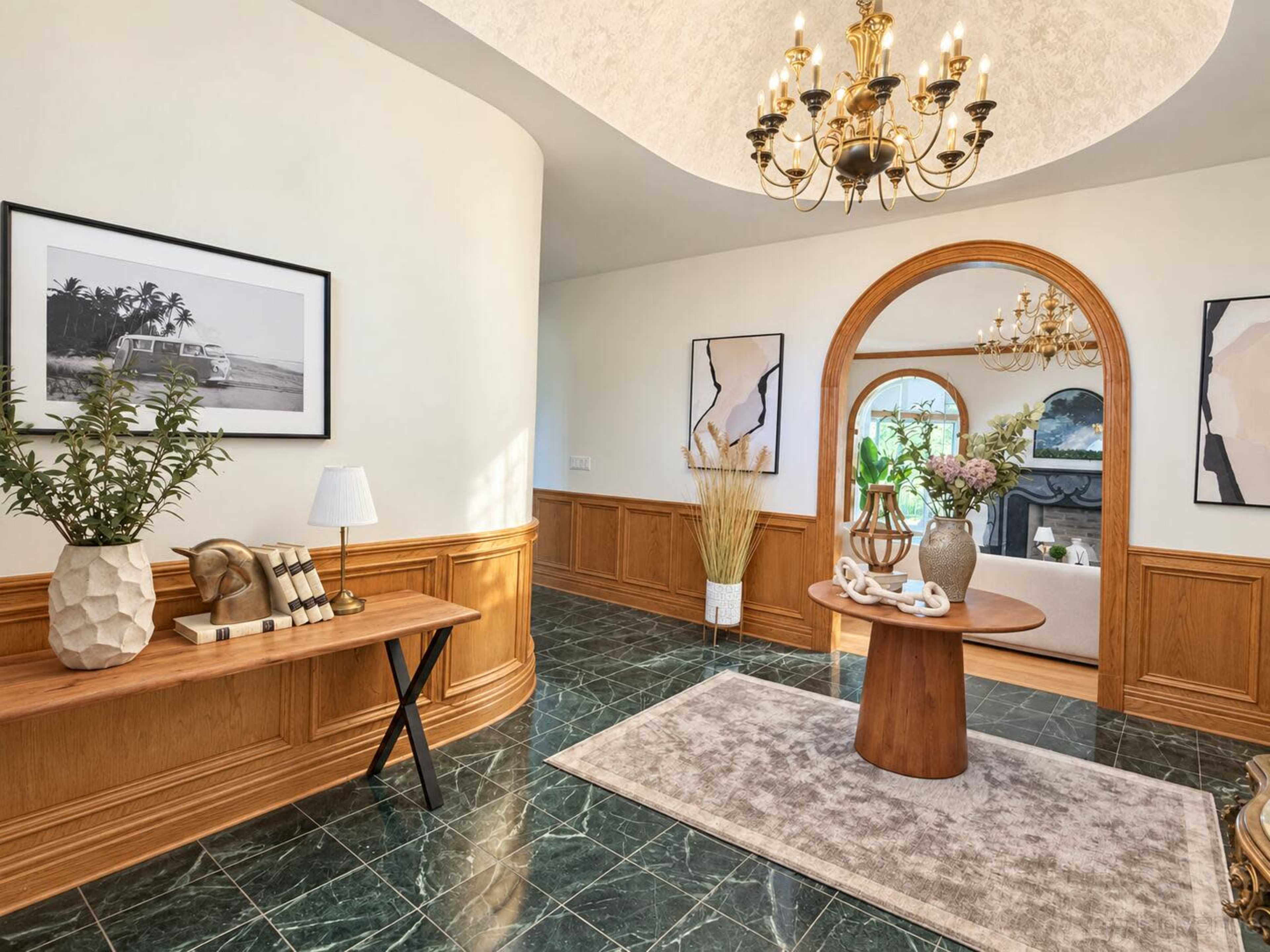 The image shows an elegantly decorated foyer featuring a dark marble floor, a round wooden table with decorative items, and framed artwork on the walls.