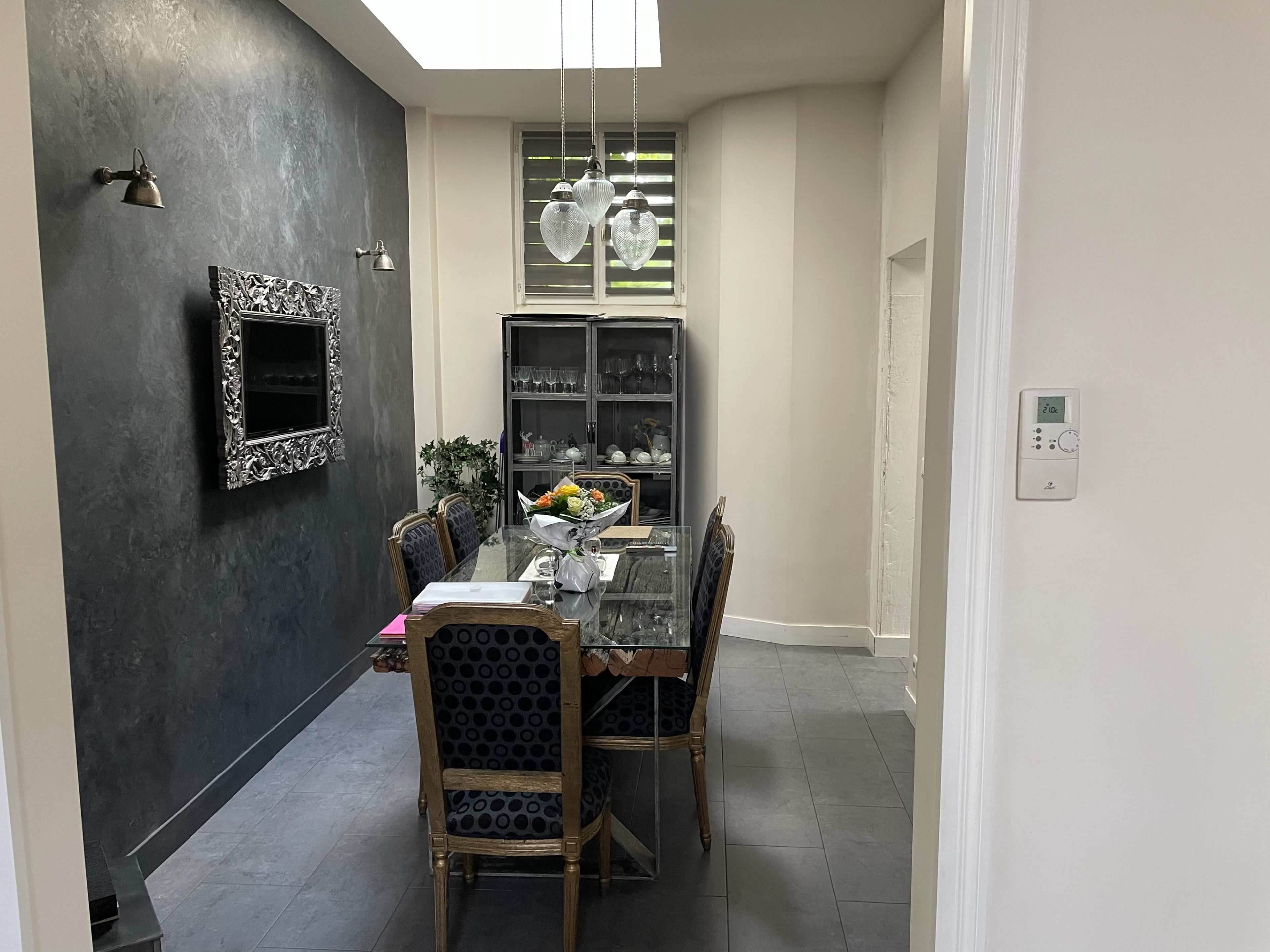 A modern dining area with a glass table surrounded by upholstered chairs, and a cabinet displaying glassware.