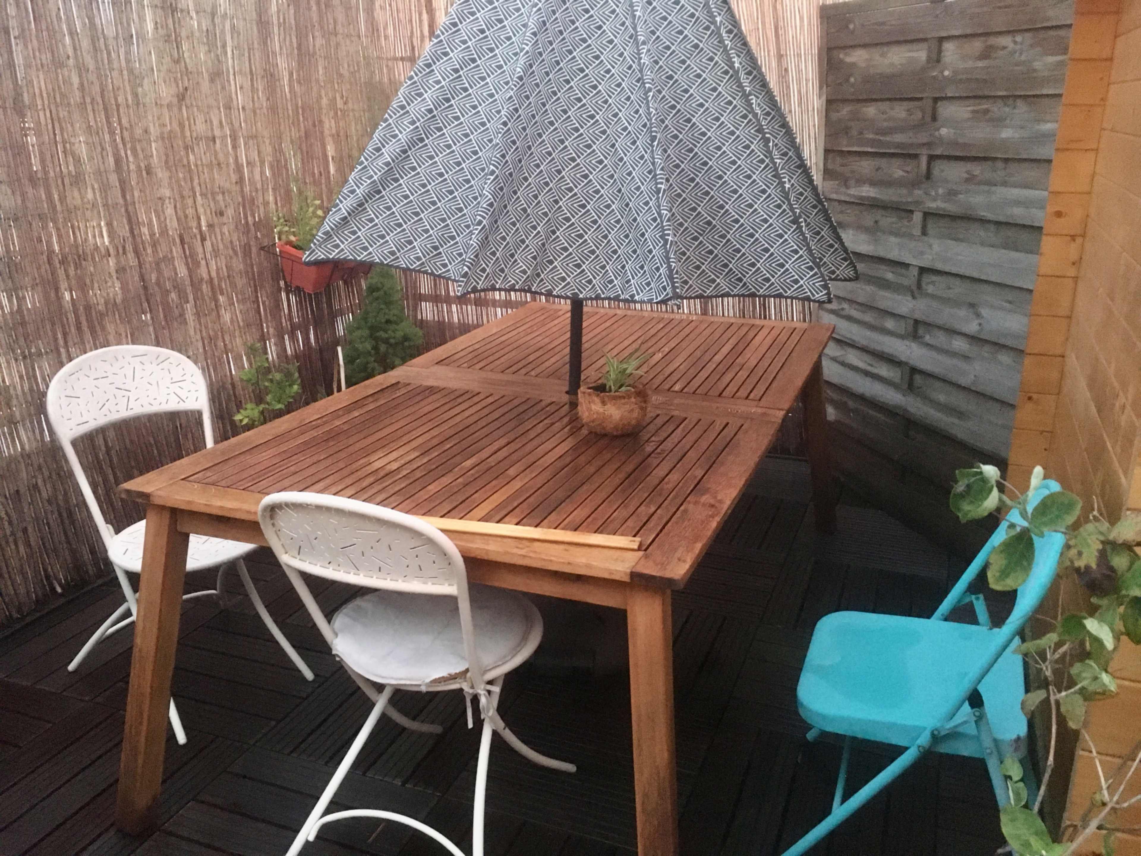 A wooden outdoor table with an umbrella stands in a small patio surrounded by wooden fences and potted plants.