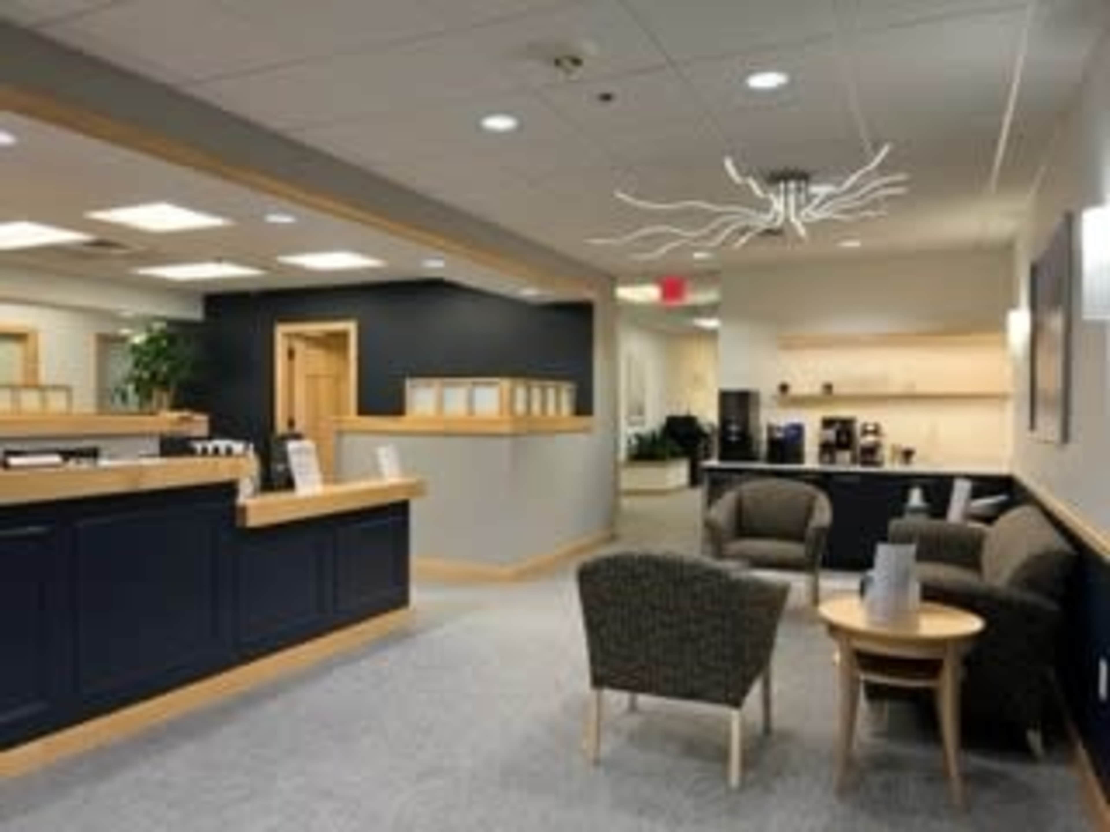 The image shows a modern reception area with a dark blue reception desk, comfortable seating, and a minimalist design featuring a light fixture.