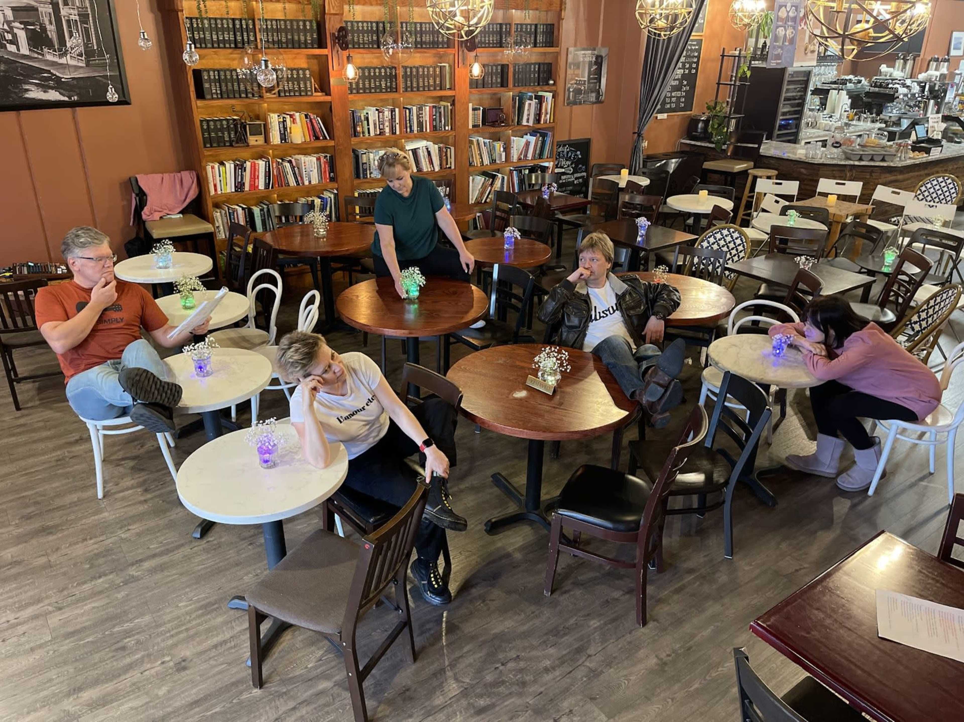 A group of five people sits at tables in a café, each with colorful drinks, while books line the walls behind them.