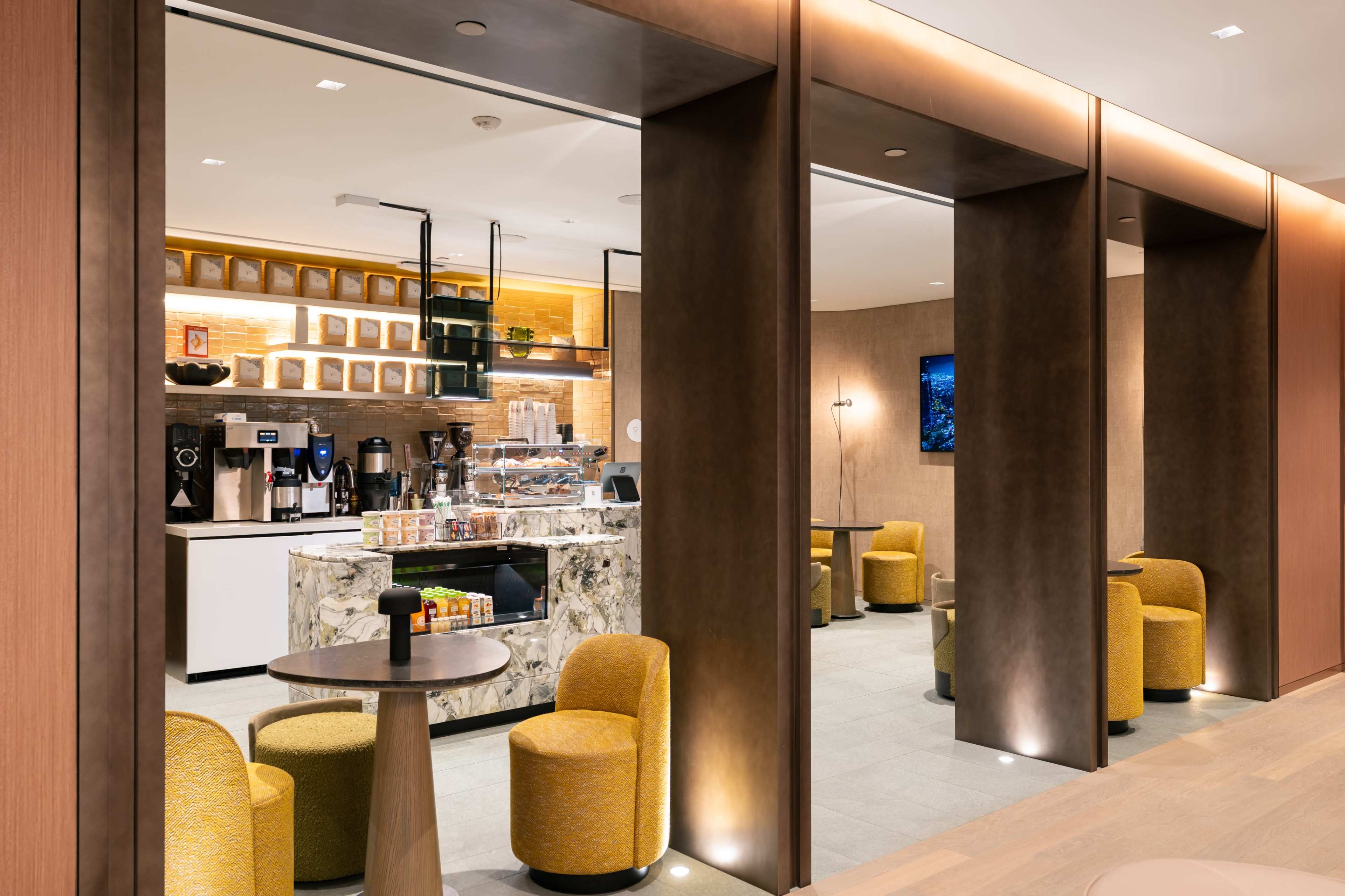 The image shows a modern café interior with a coffee bar featuring a marble countertop and several seating areas with circular tables and yellow upholstered chairs.