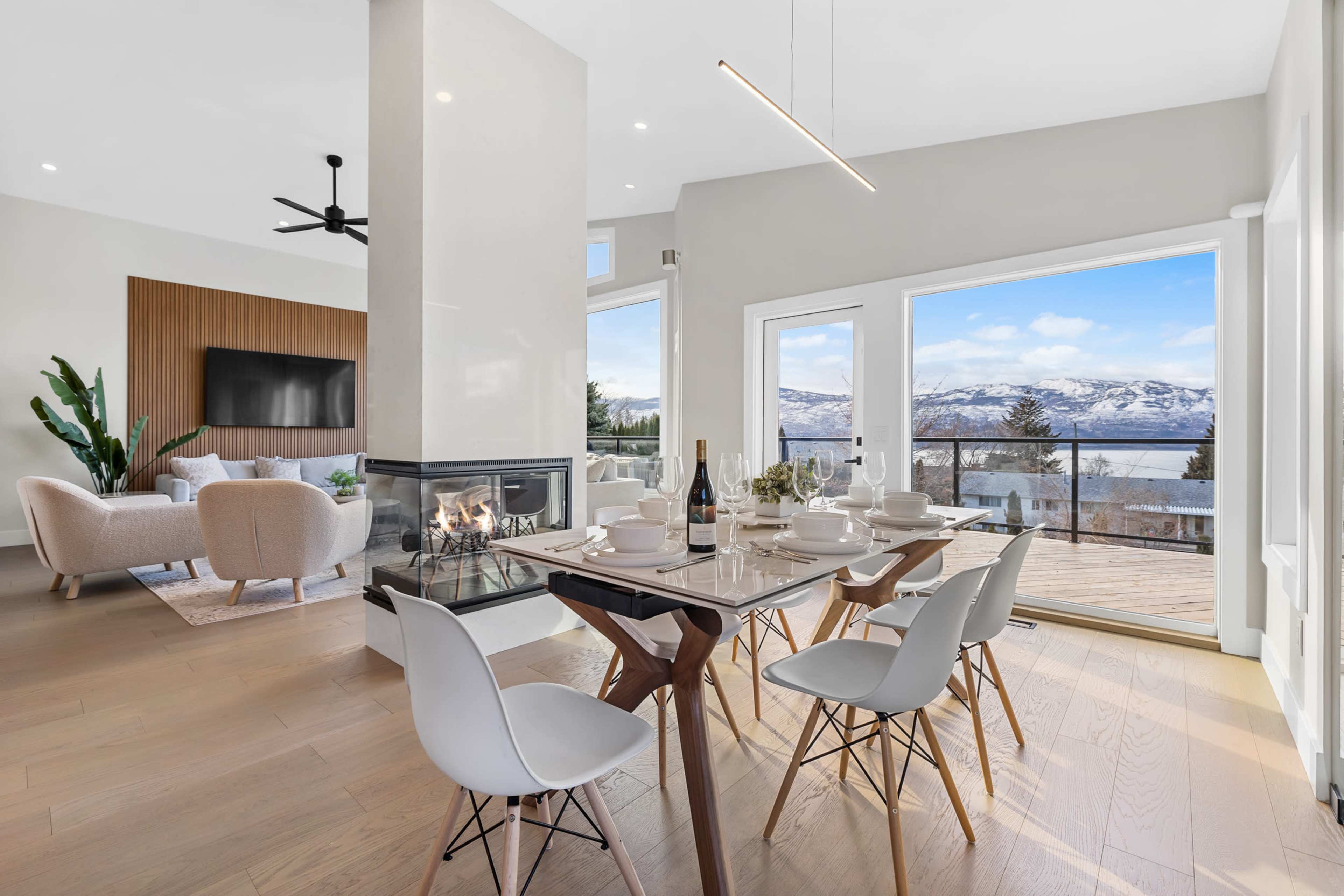 A modern dining area features a glass table set for six, with a view of a lake and mountains through large windows.