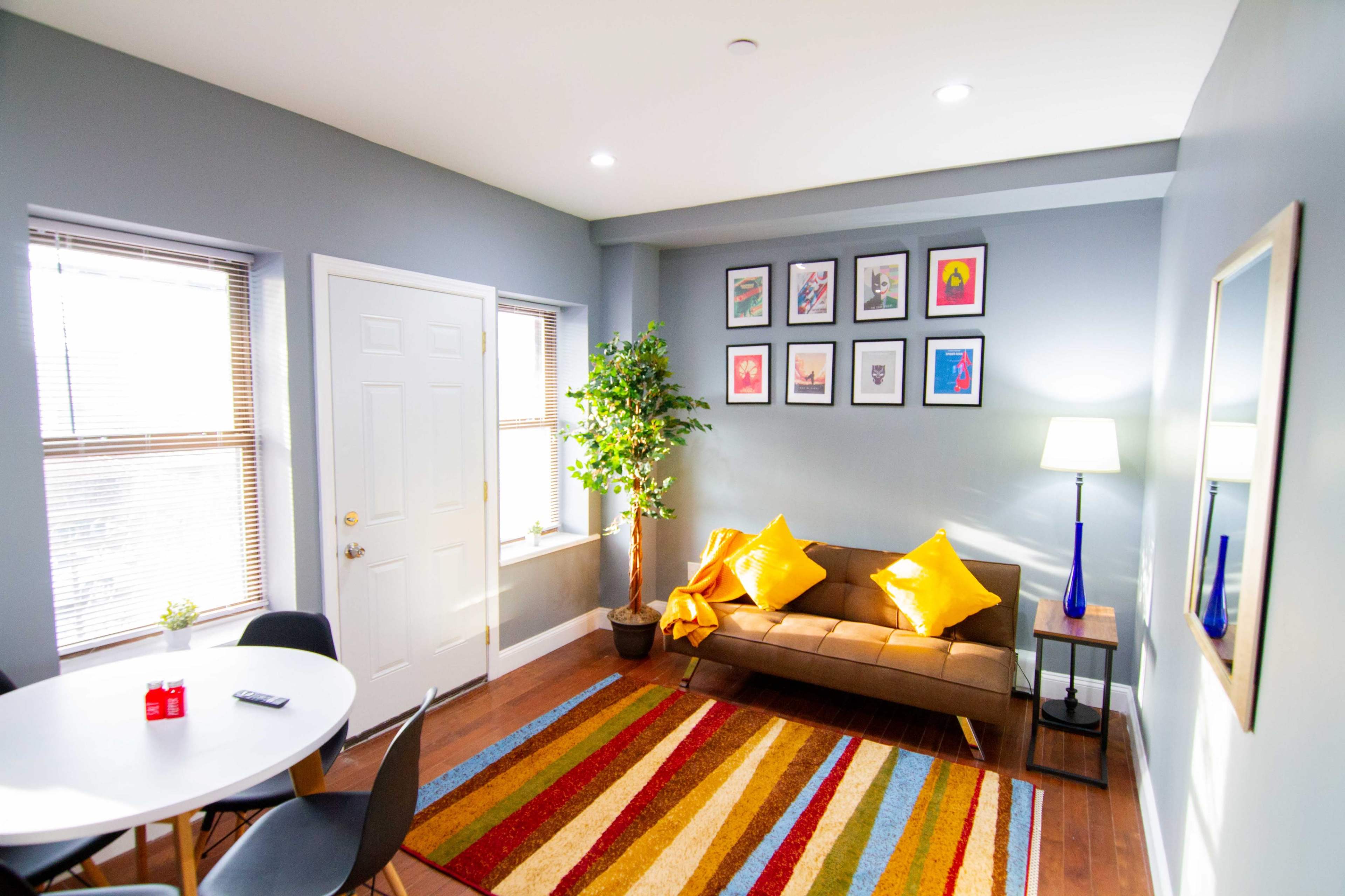 A living room features a brown couch with yellow cushions, a round dining table, multicolored striped rug, and framed artwork on the wall.
