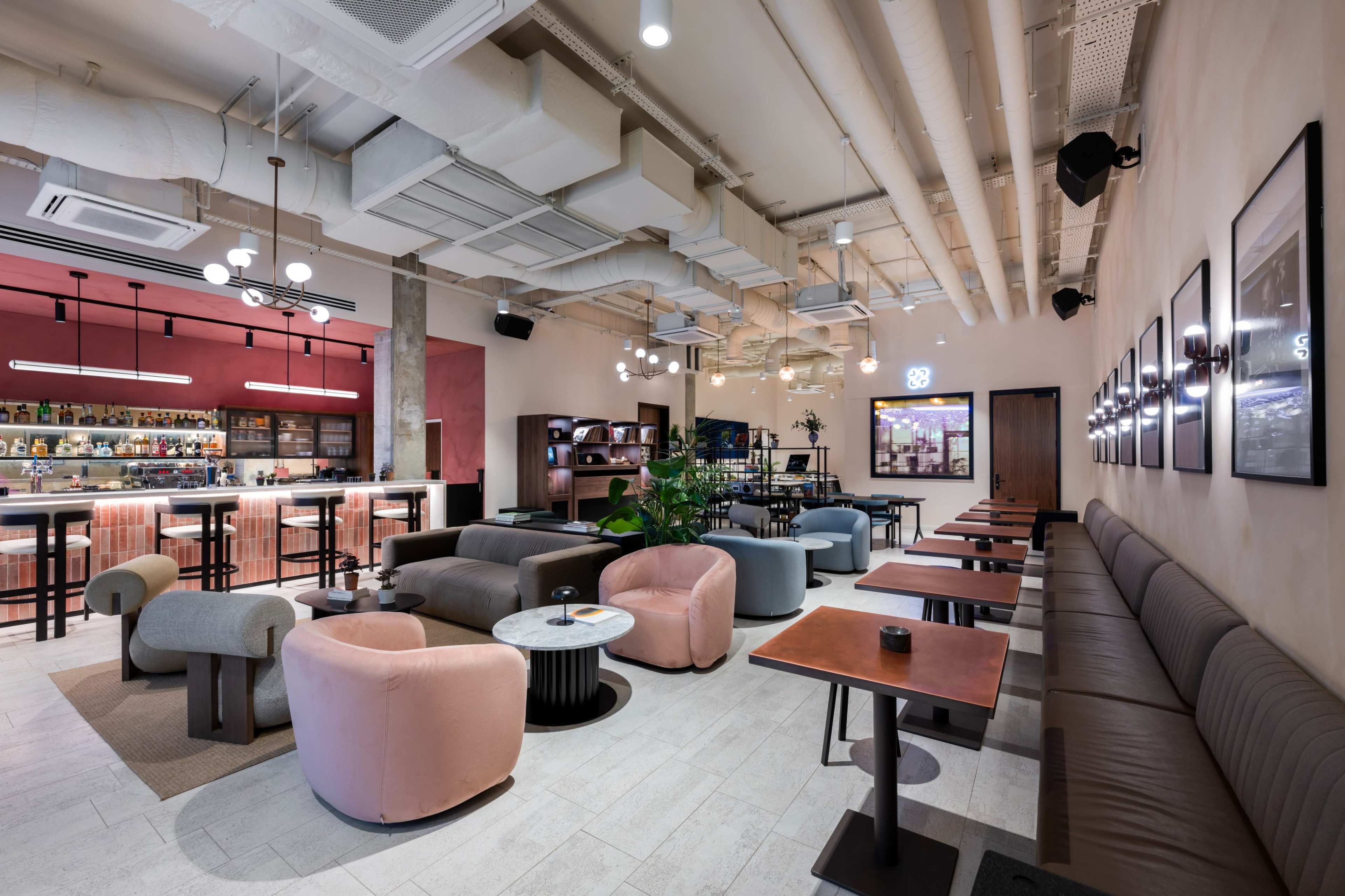 The image shows a modern, spacious lounge with a bar area, featuring various seating arrangements and a warm color palette.