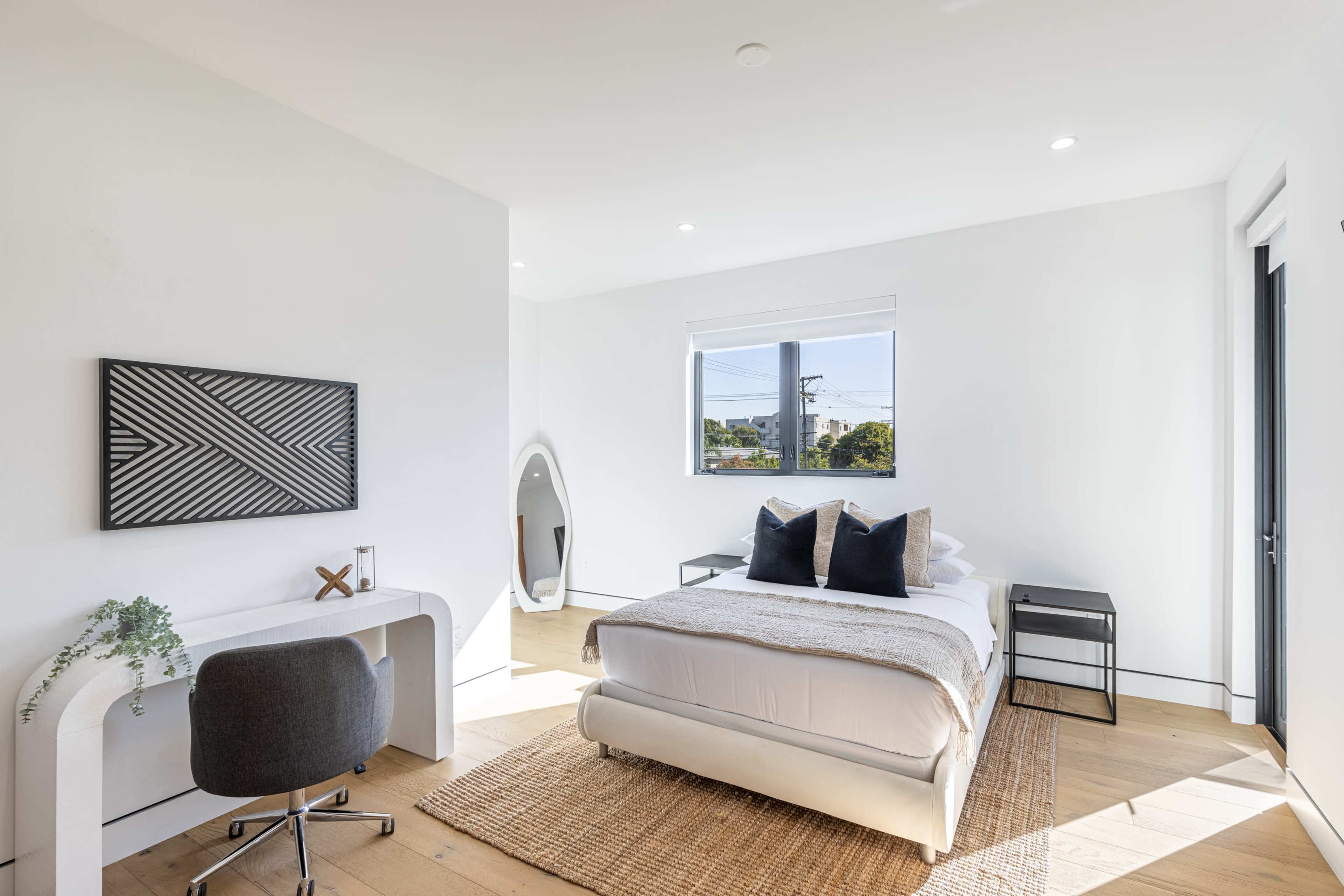 A bright bedroom features a bed with white linens, a desk, a chair, and a round mirror against the wall, set on light wood flooring.
