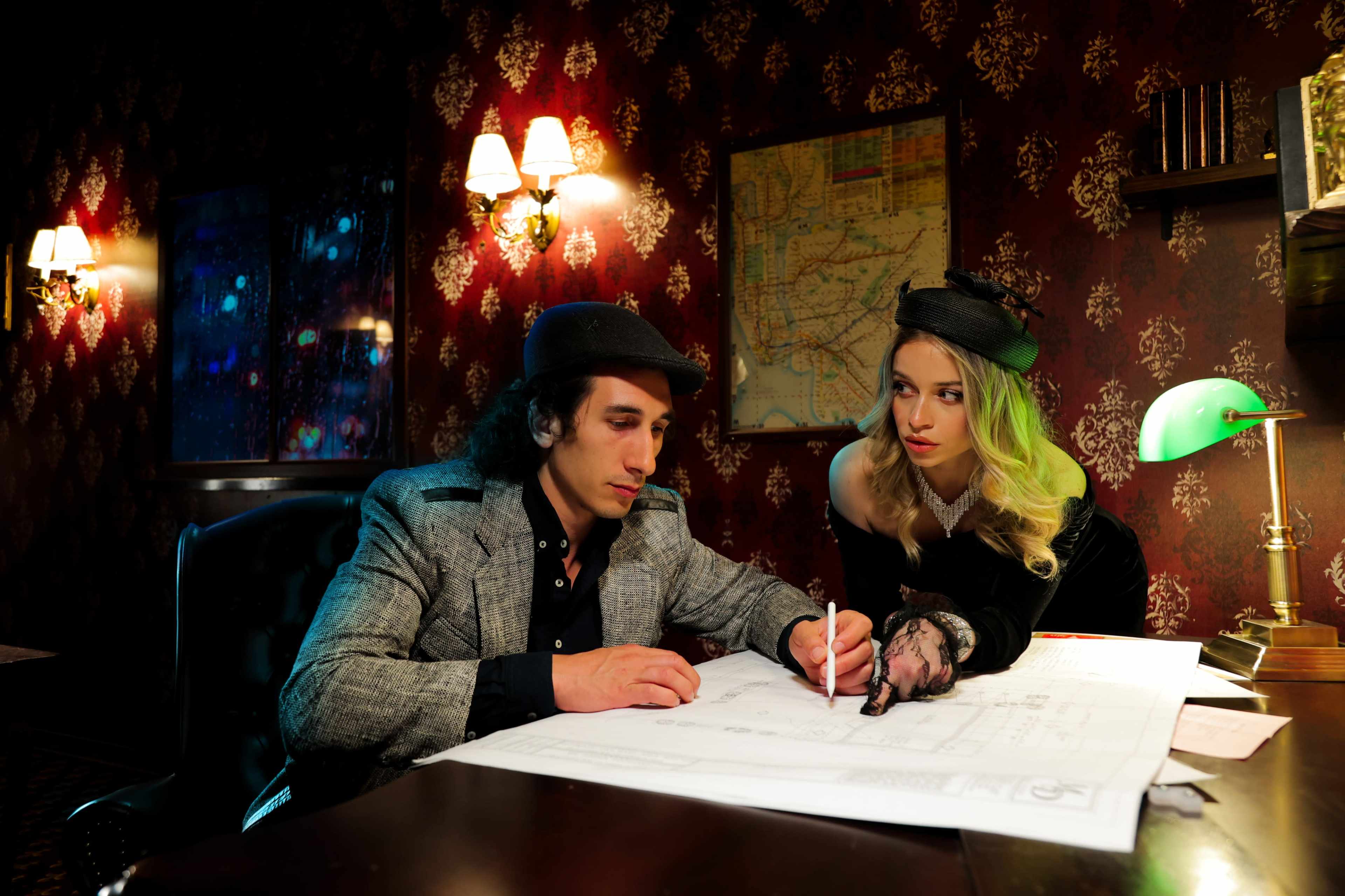 A man and woman examine architectural plans on a table in a dimly lit, vintage-style room with wallpaper and antique lamps.