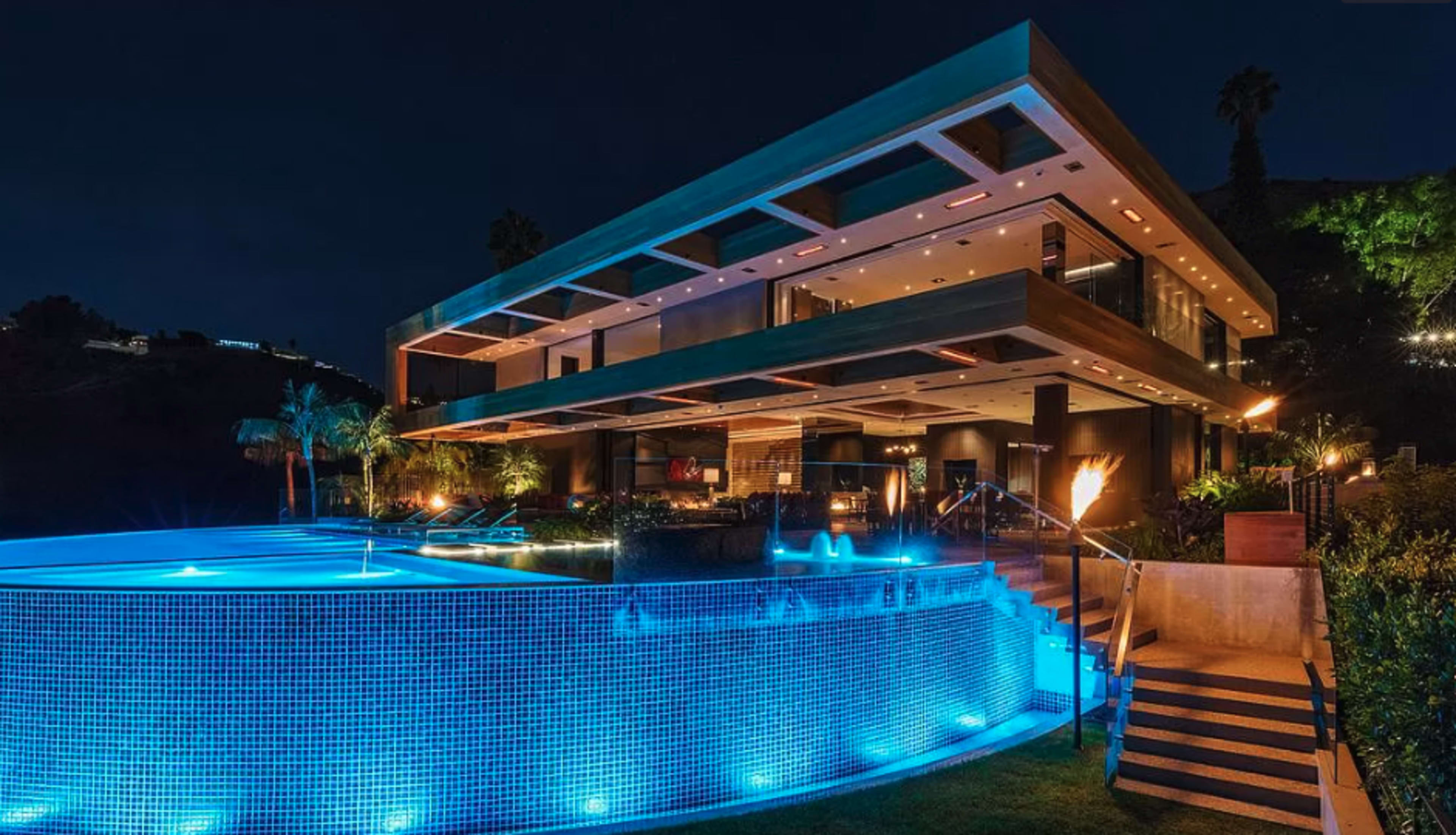 The image shows a modern, multi-level house illuminated at night, with a large pool featuring blue lighting in the foreground.