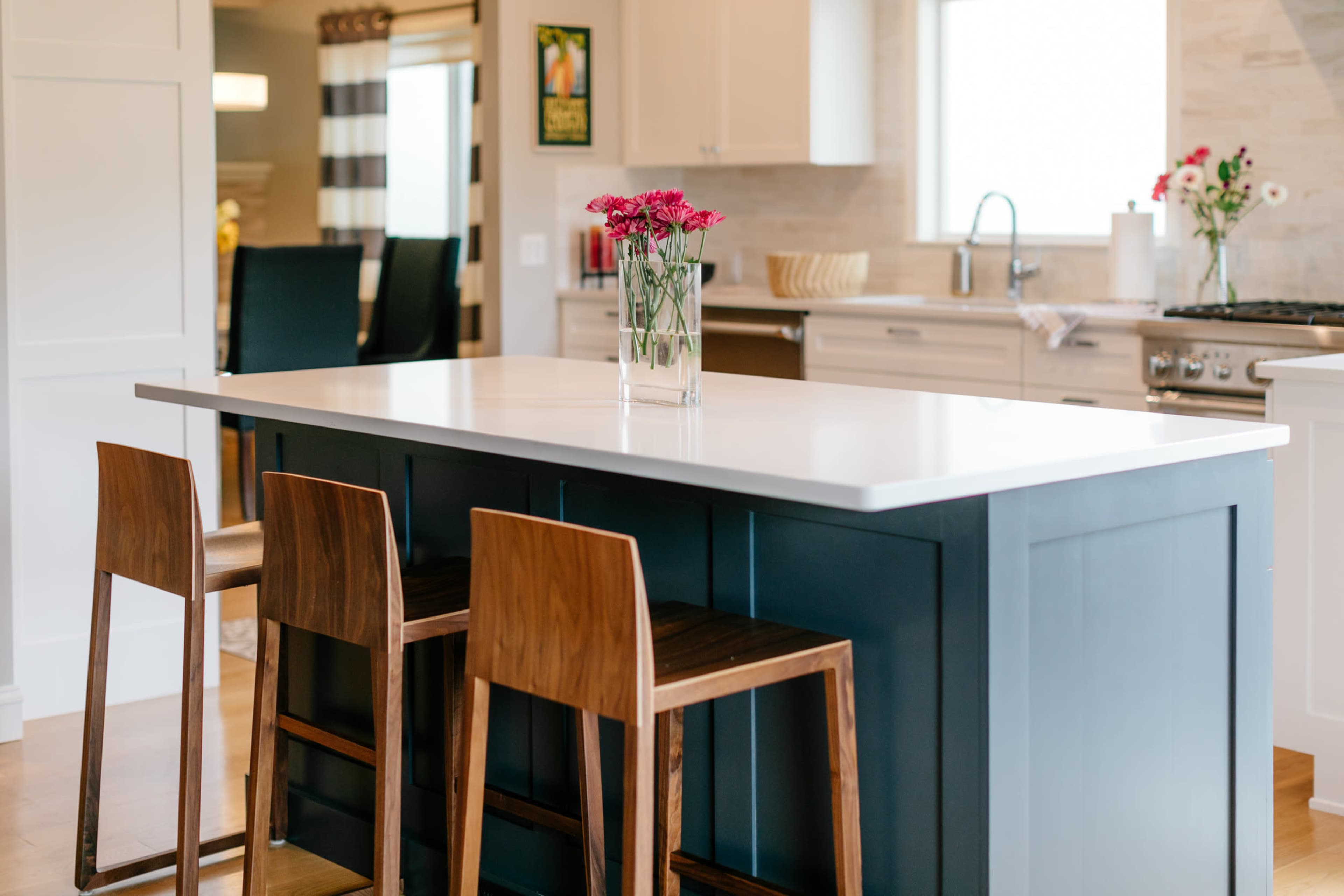 A modern kitchen features a central island with a white countertop and three wooden bar stools, adorned with a vase of pink flowers.