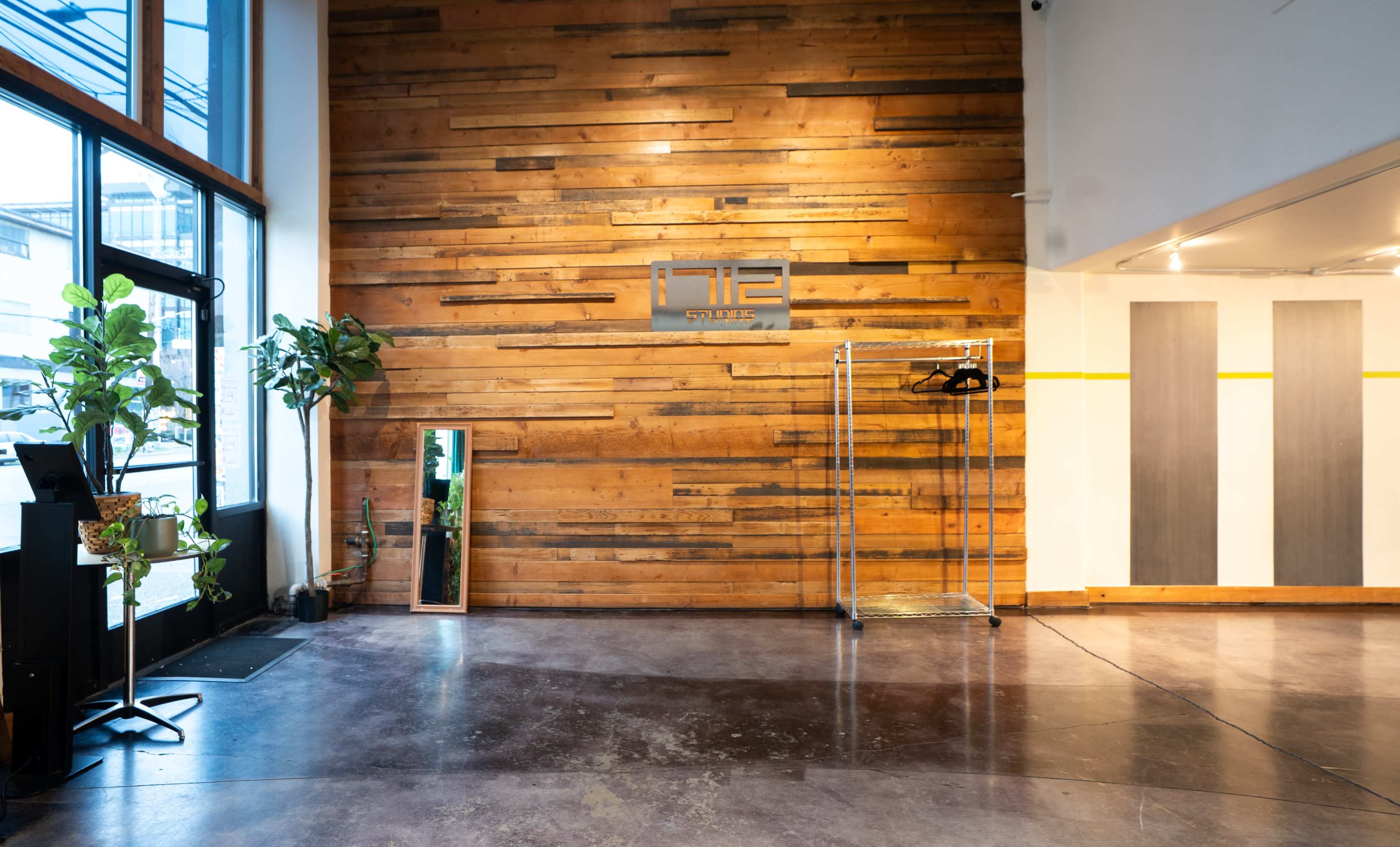 The image shows an interior space with a wooden accent wall, a mirror, a clothing rack, and a doorway leading outside.
