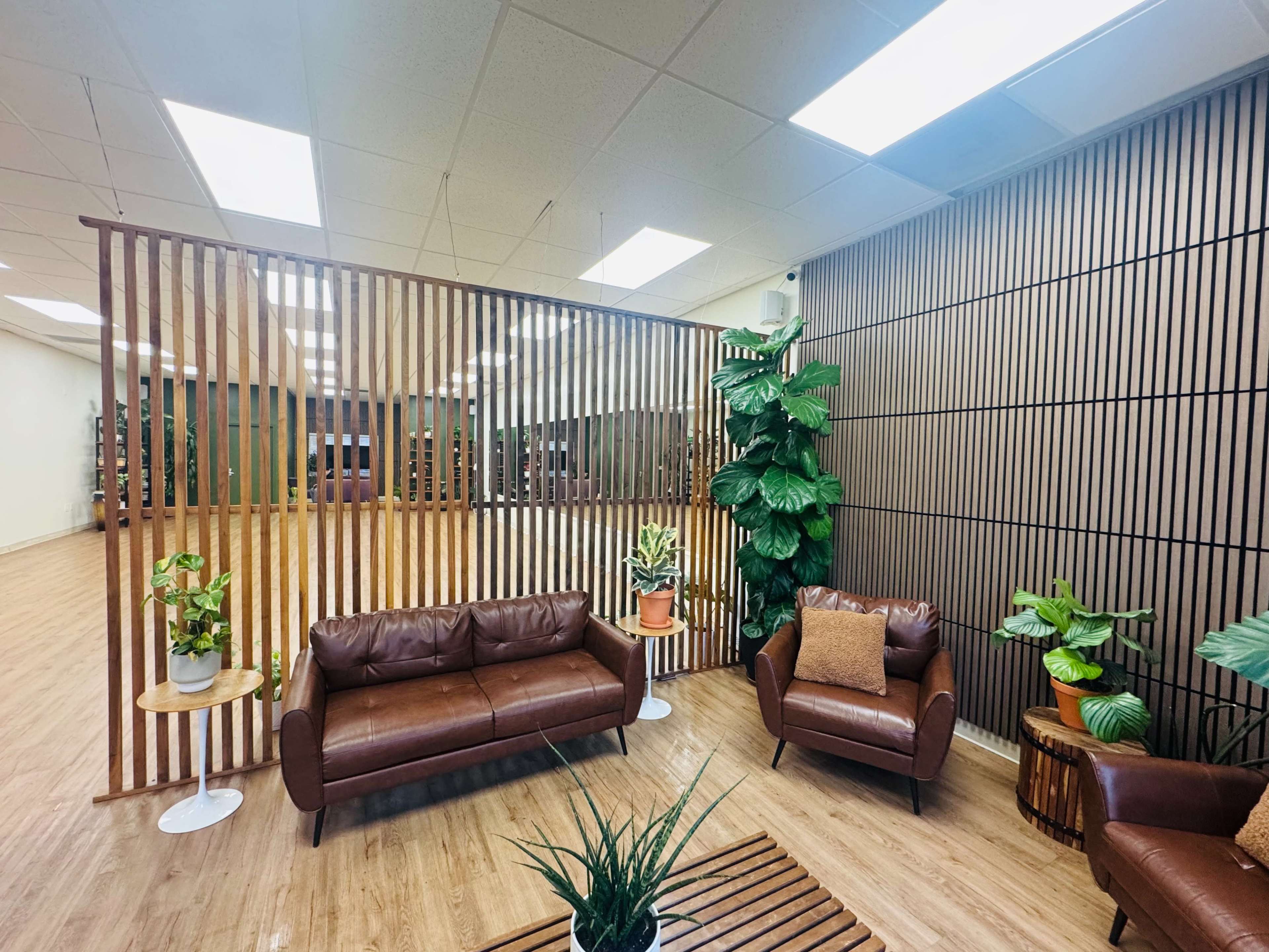 The image shows a modern waiting area with two brown leather sofas, a wooden slatted partition, and a variety of potted plants.