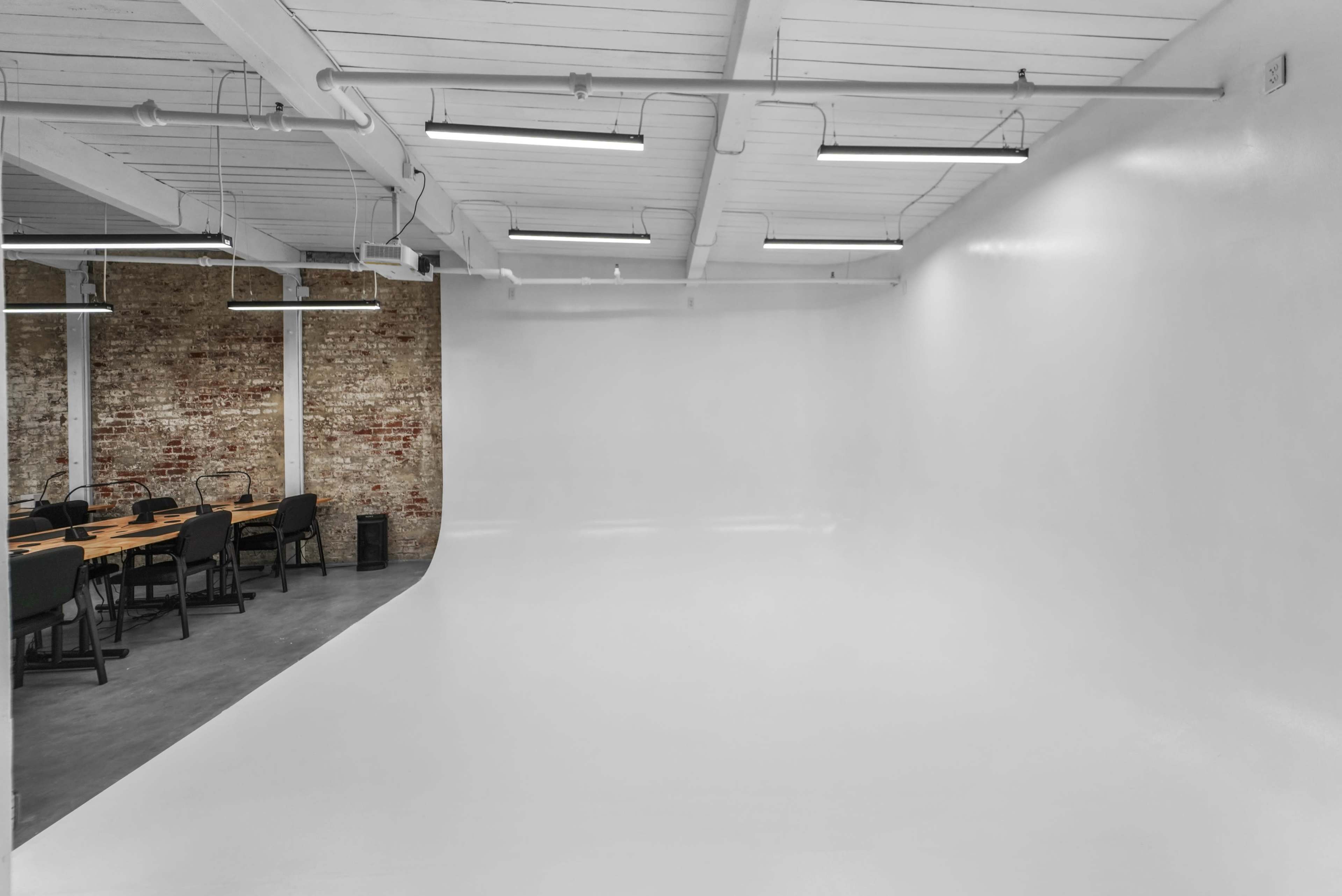 The image shows a spacious, minimalistic studio with a white backdrop and a corner meeting area featuring a long table and black chairs.