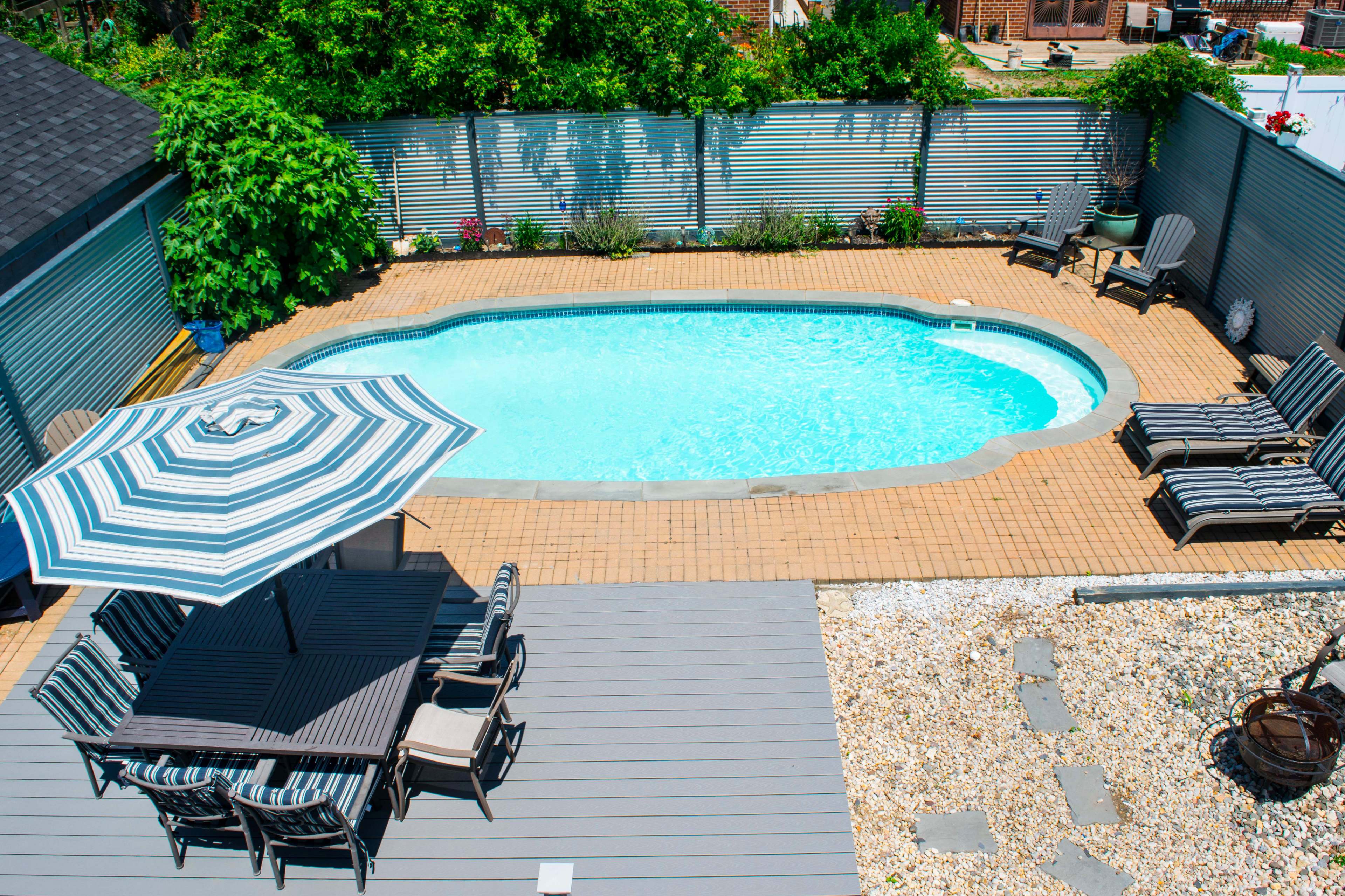 The image shows a rectangular swimming pool surrounded by a patio with lounge chairs and a dining area shaded by a striped umbrella.