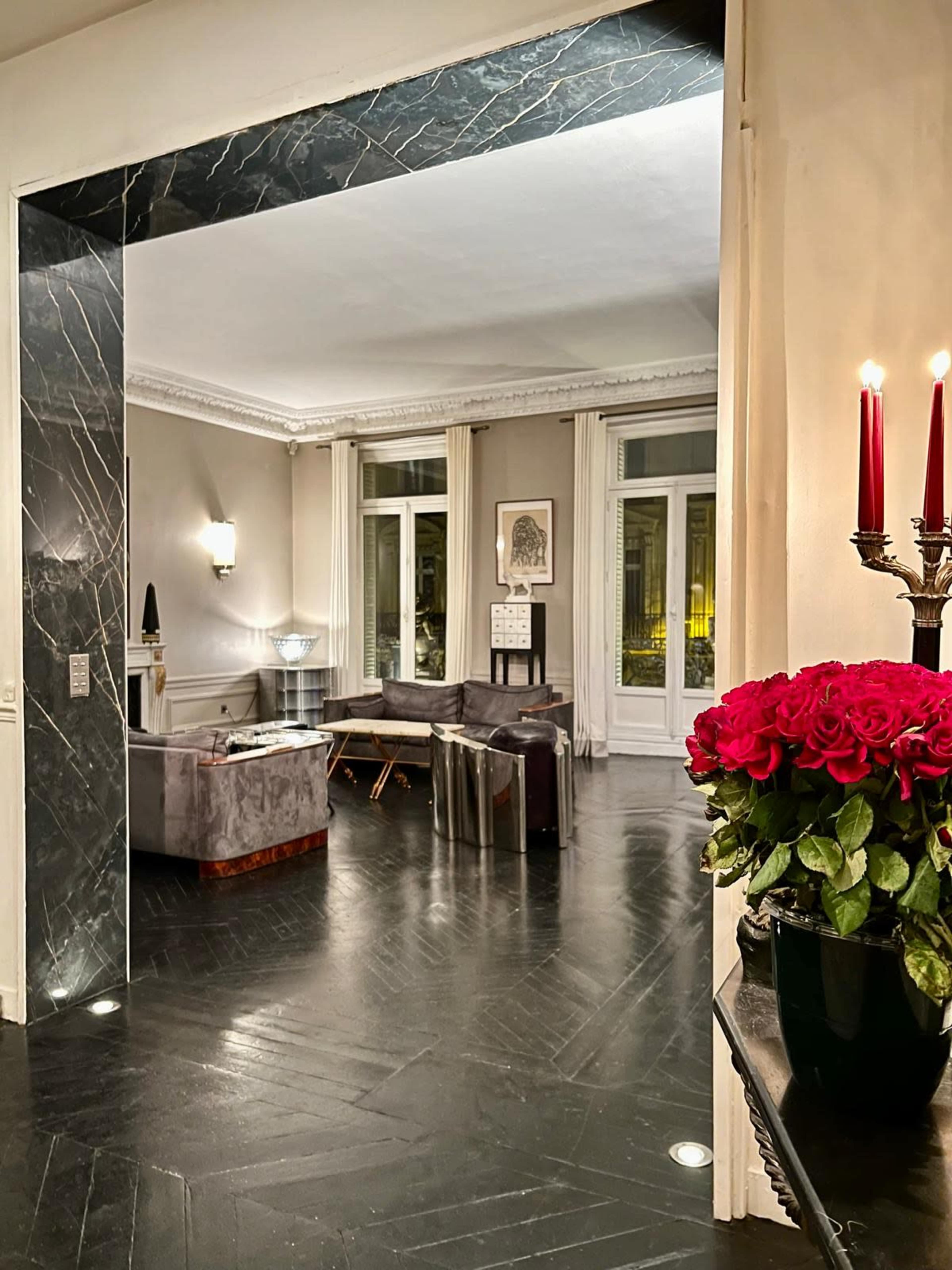 The image depicts a well-decorated living room with large windows, contemporary furniture, and a vase of red roses on a nearby table.