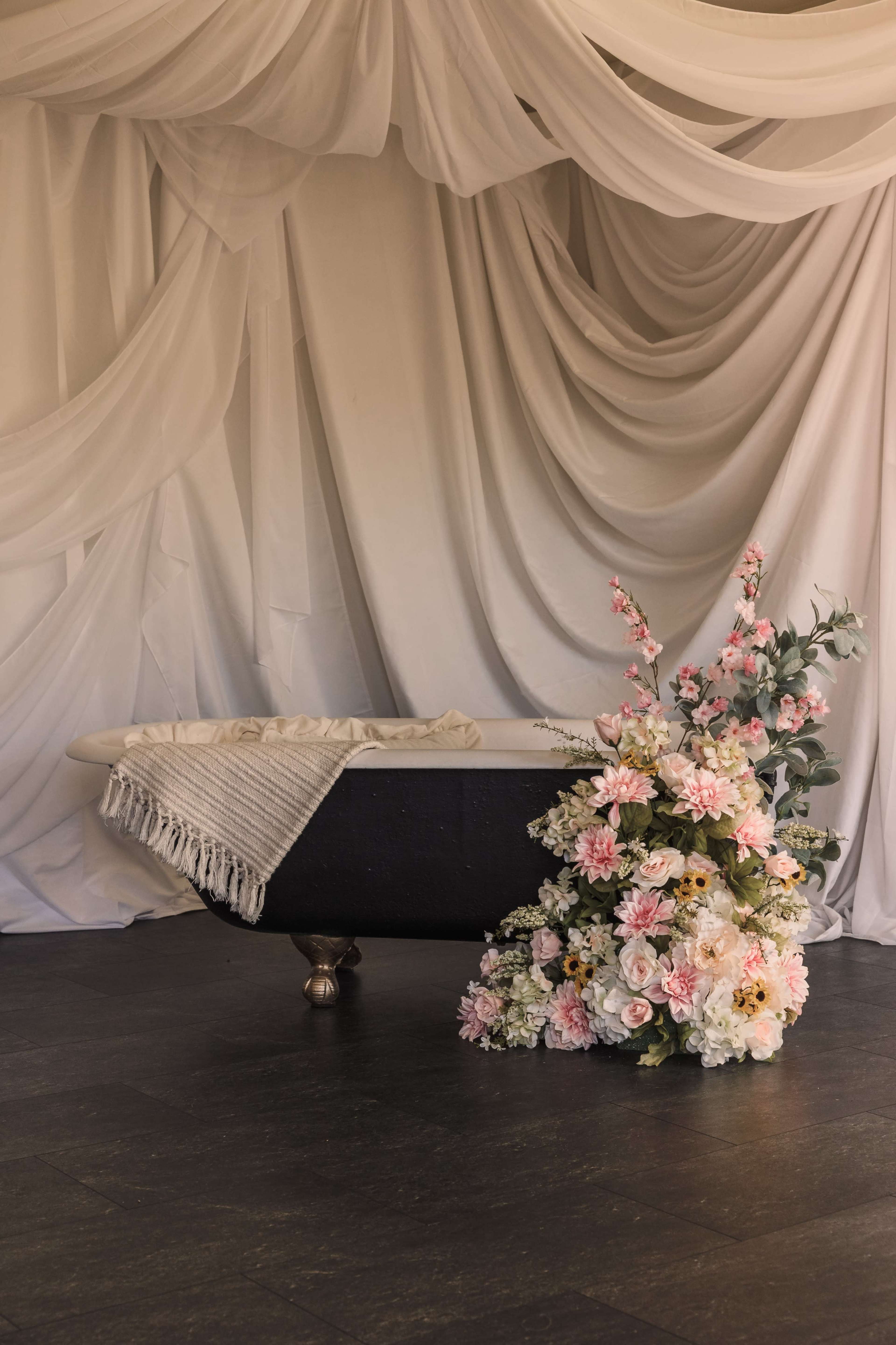 A vintage-style bathtub is positioned against a backdrop of draped white fabric, accompanied by a large arrangement of colorful artificial flowers.