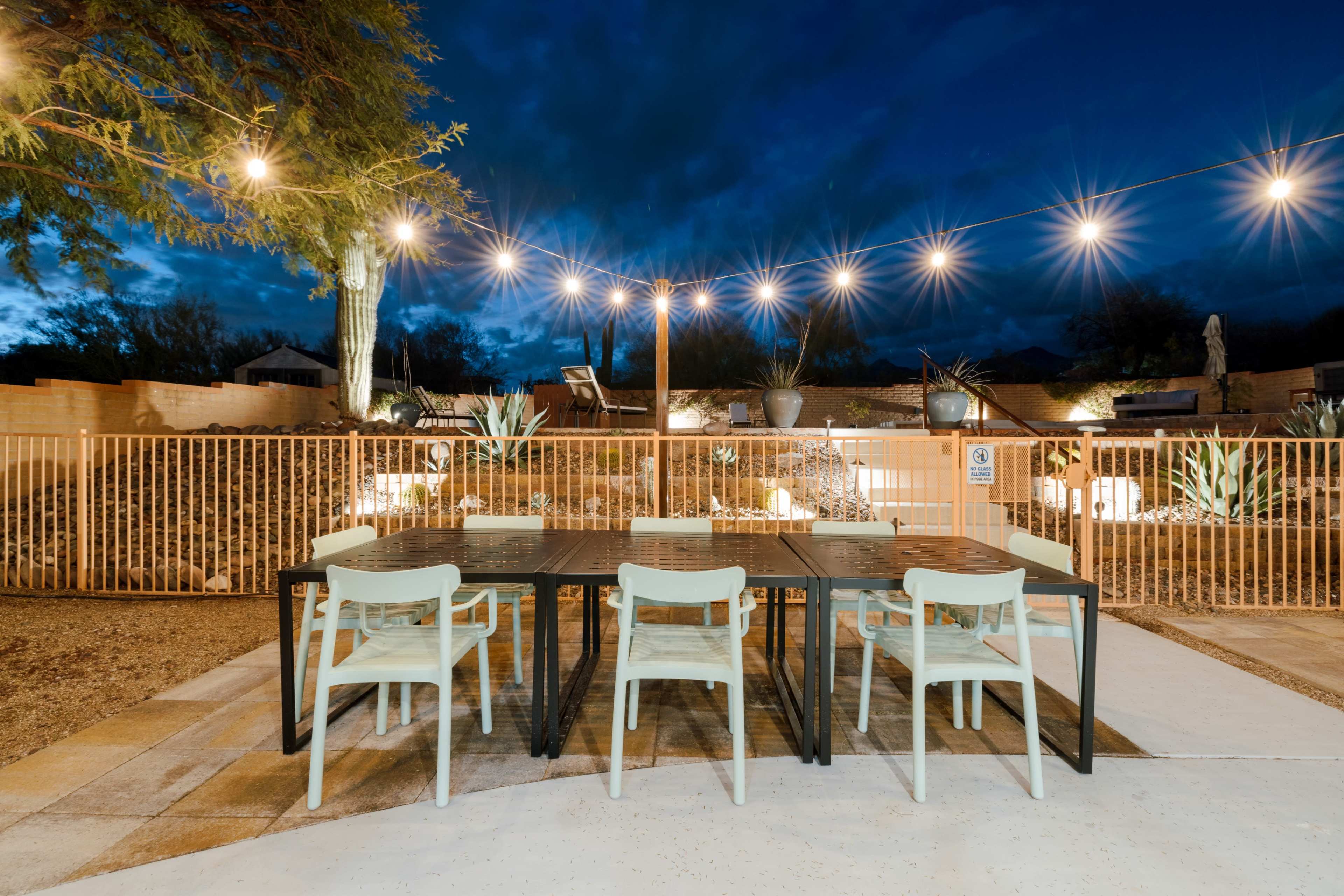 A dining area with a large table and chairs is illuminated by string lights against a nighttime backdrop of clouds and desert landscaping.