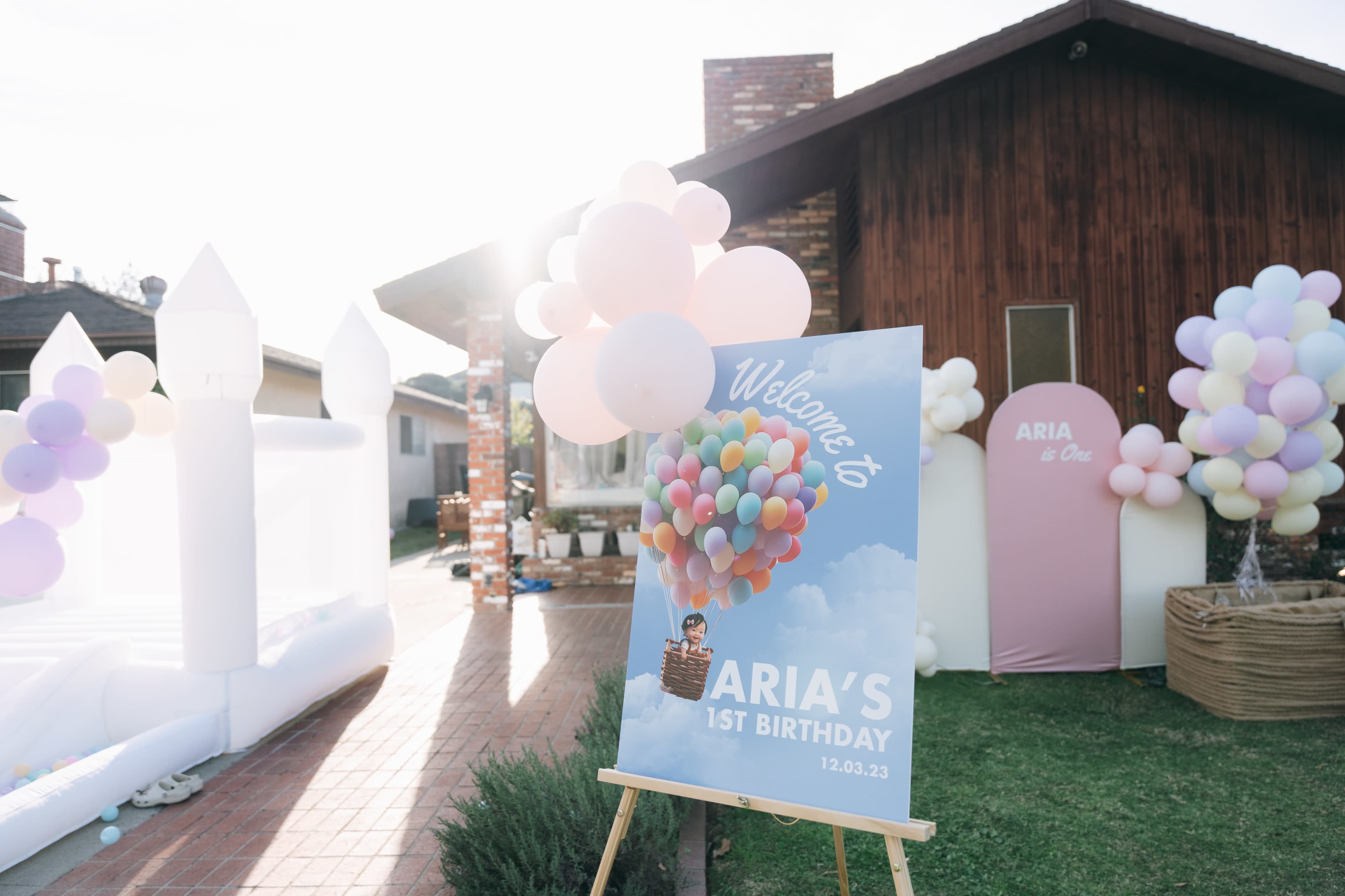 A colorful sign welcomes guests to a celebration for a child's first birthday, featuring balloon decorations and a backdrop of a house with a bounce house nearby.