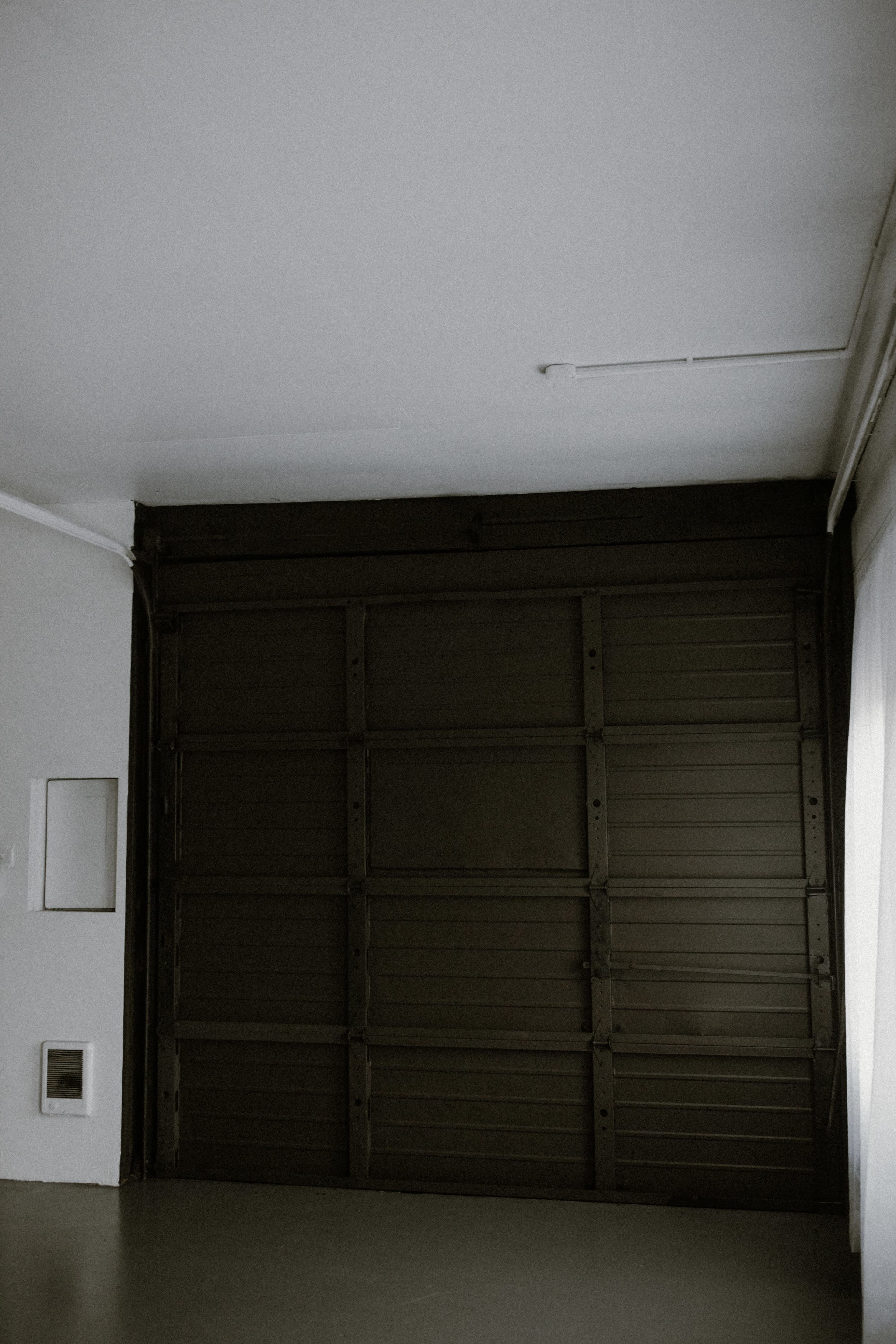 A minimalistic interior space with a large closed metal garage door and a white wall with a small square opening.