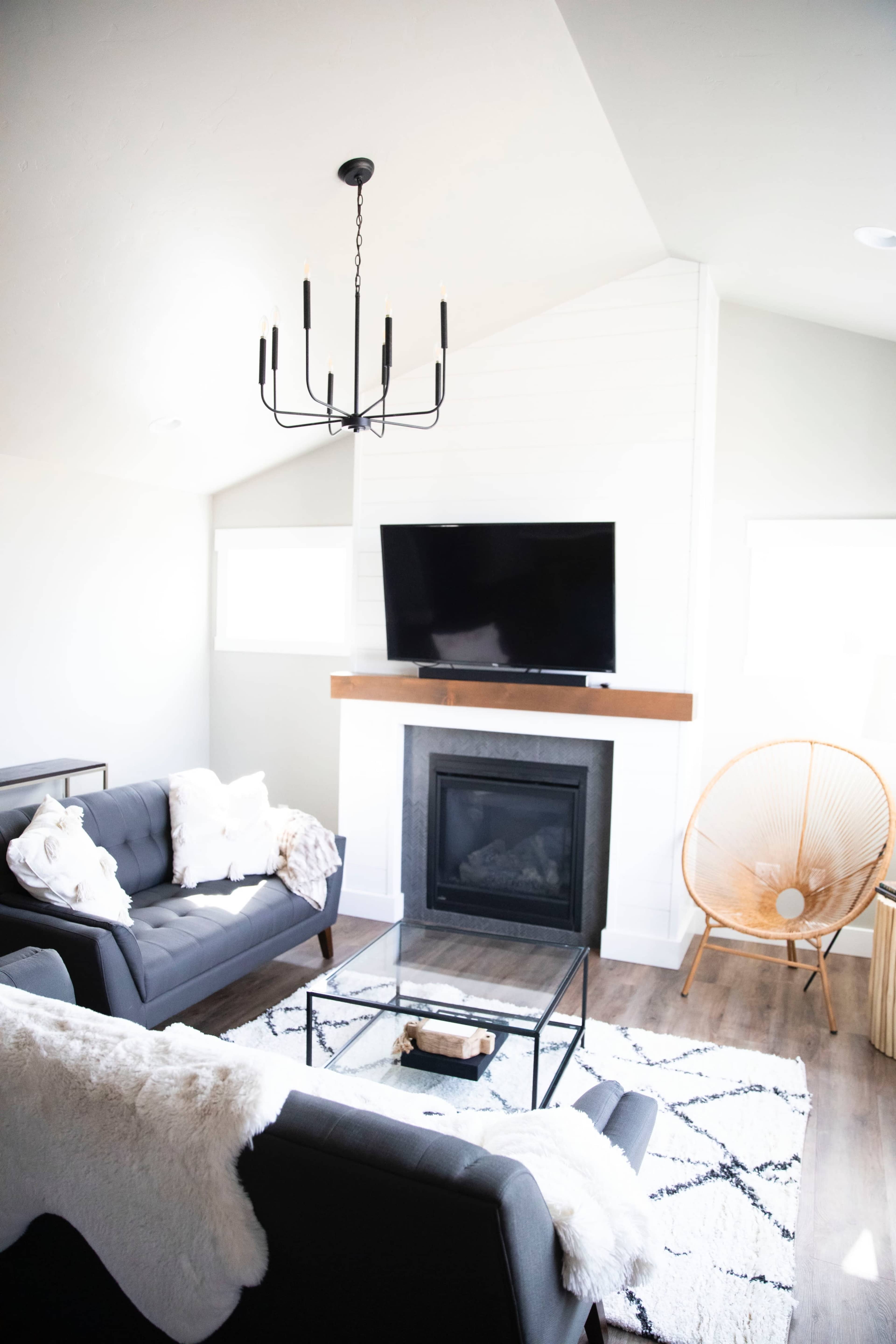 A modern living room featuring a fireplace with a mounted TV above it, a black sofa, an armchair, and a glass coffee table on a patterned rug.
