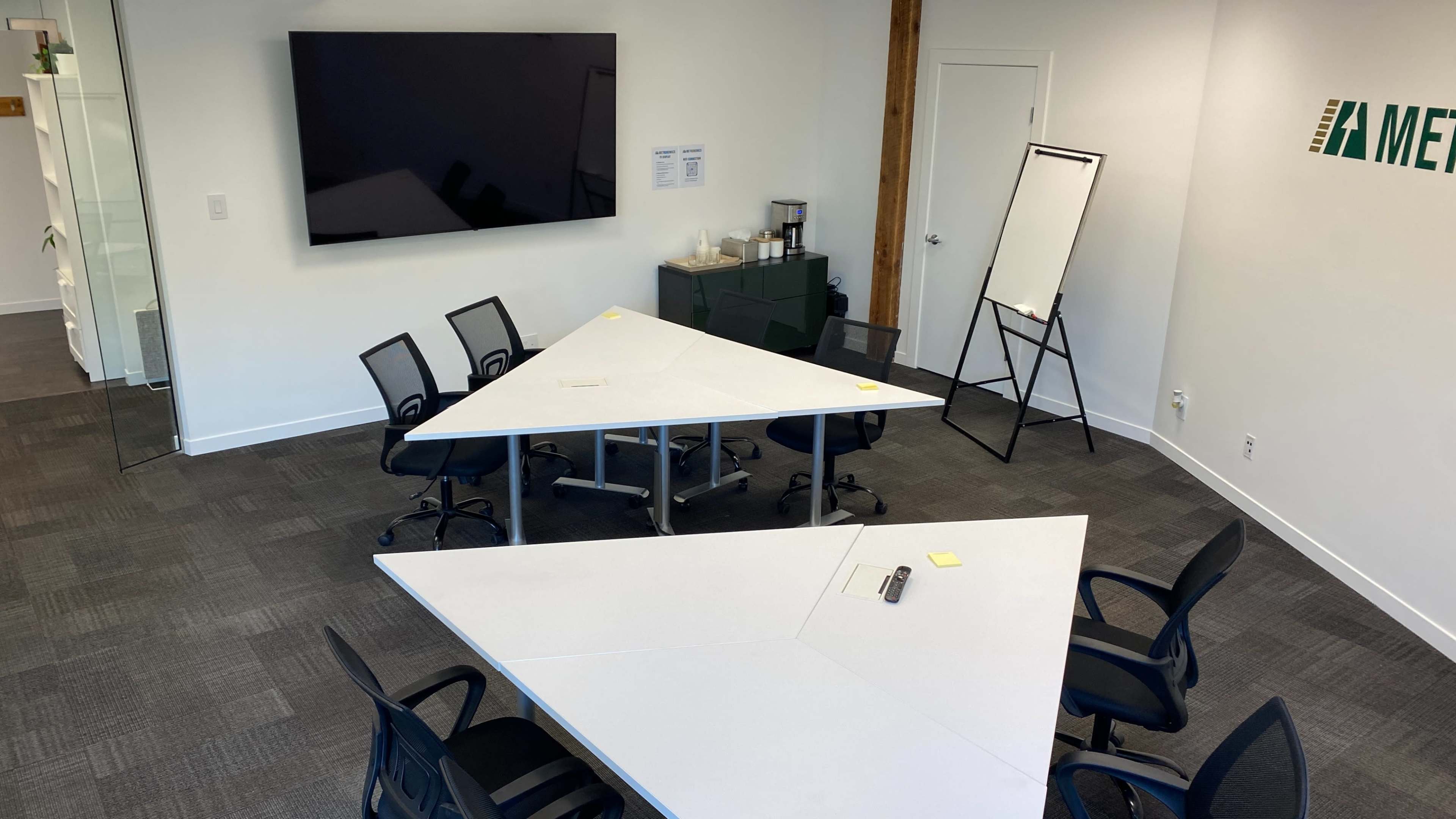 A modern conference room features two triangular tables with six chairs, a large wall-mounted screen, and a whiteboard.