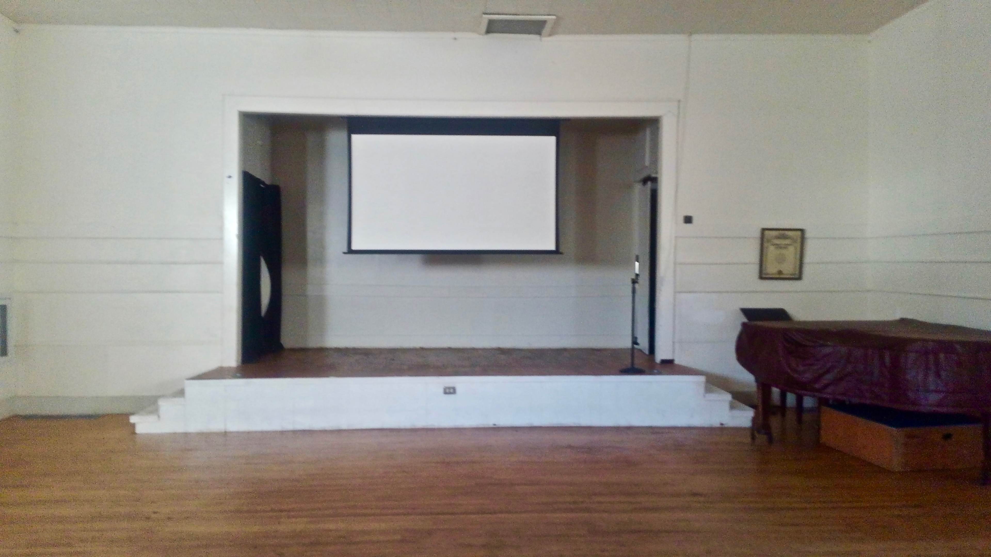 The image shows a simple stage with a projector screen and a microphone stand in a sparsely decorated room with wooden flooring.