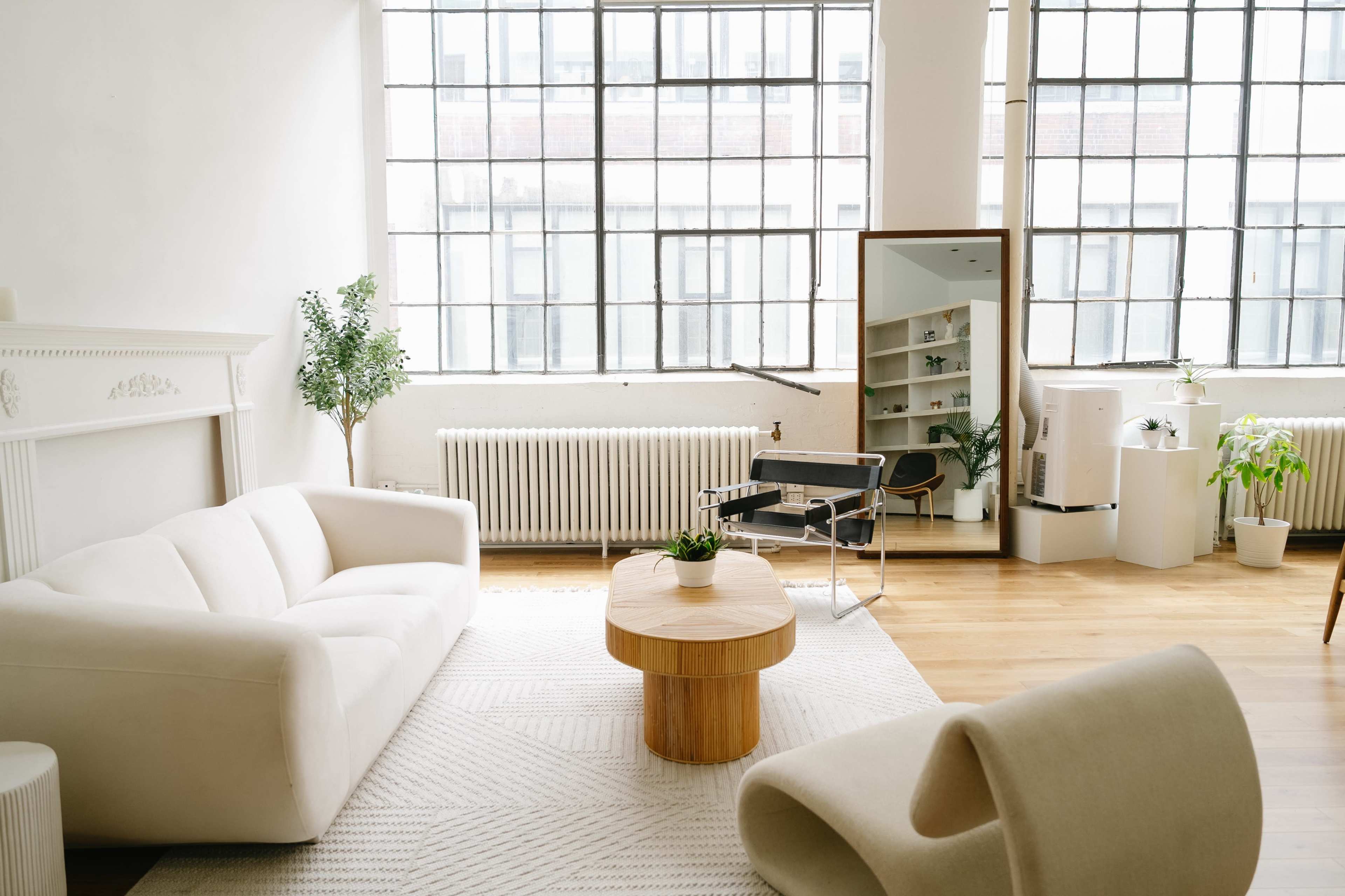 The image shows a bright, airy living space with large windows, a light-colored sofa, a wooden coffee table, and a mirror reflecting shelves with plants.