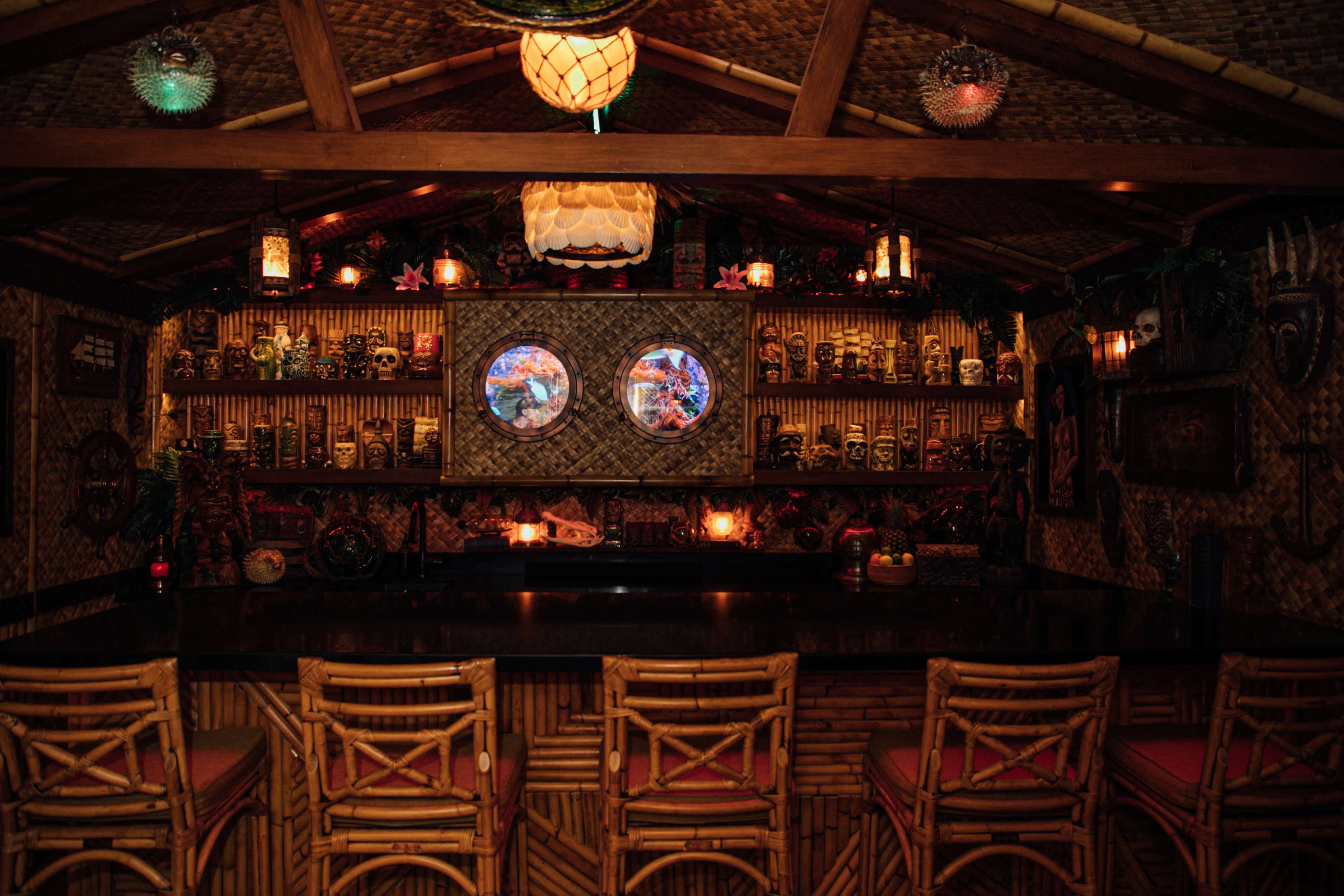 A tiki-themed bar with a bamboo structure, decorated shelves lined with various masks and skulls, and circular windows illuminated by colorful lights.