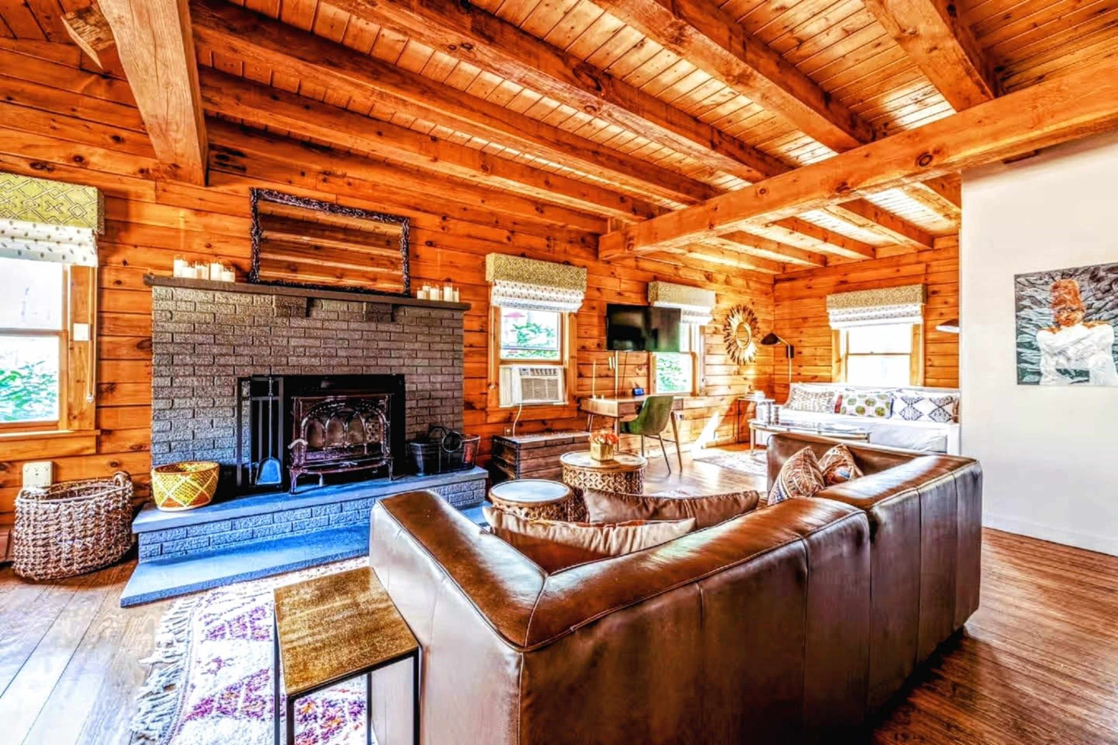 The image shows a cozy wooden interior of a living room featuring a stone fireplace, leather couch, and a dining table near a window.
