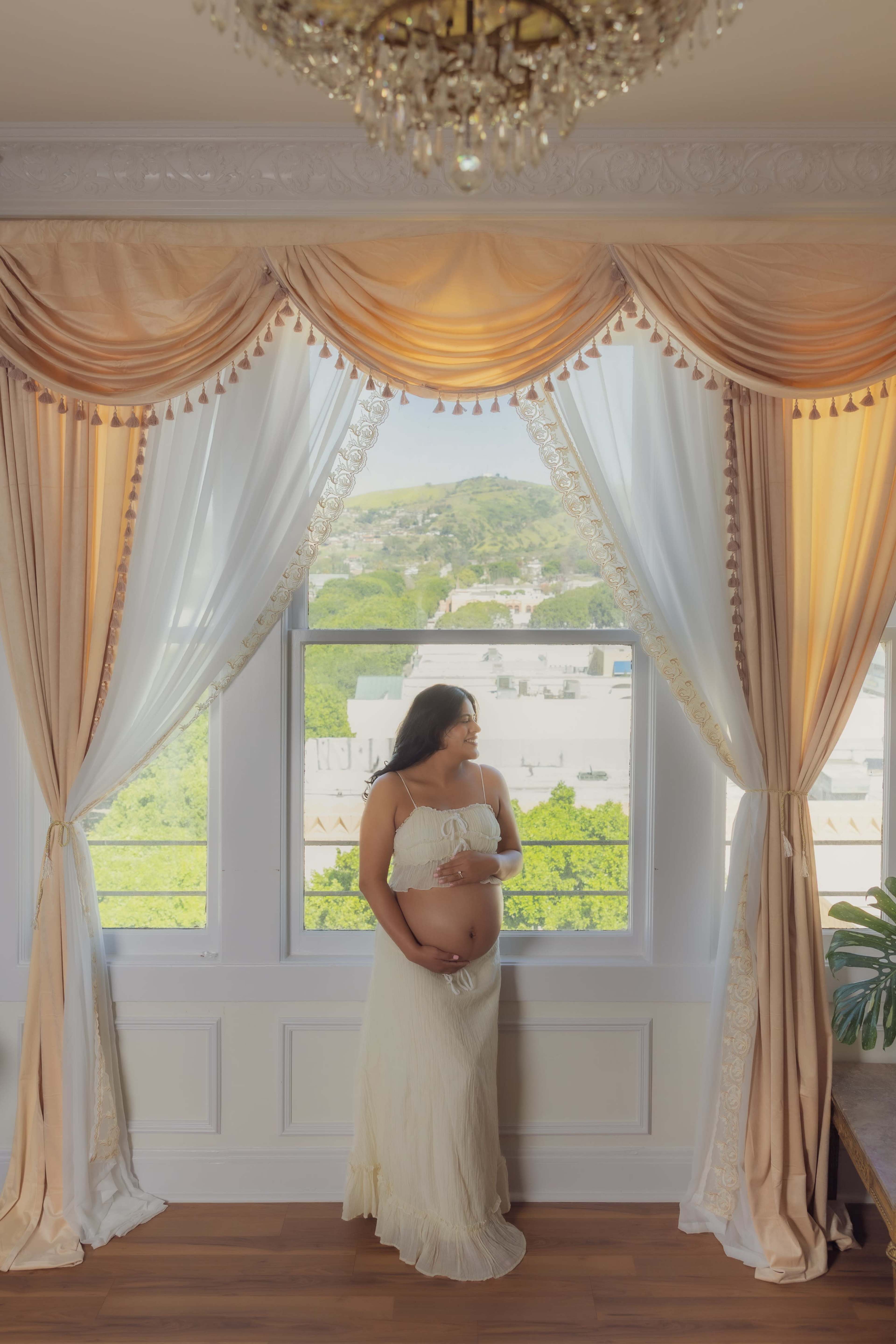 A pregnant woman wearing a white dress stands by a window adorned with elegant curtains.