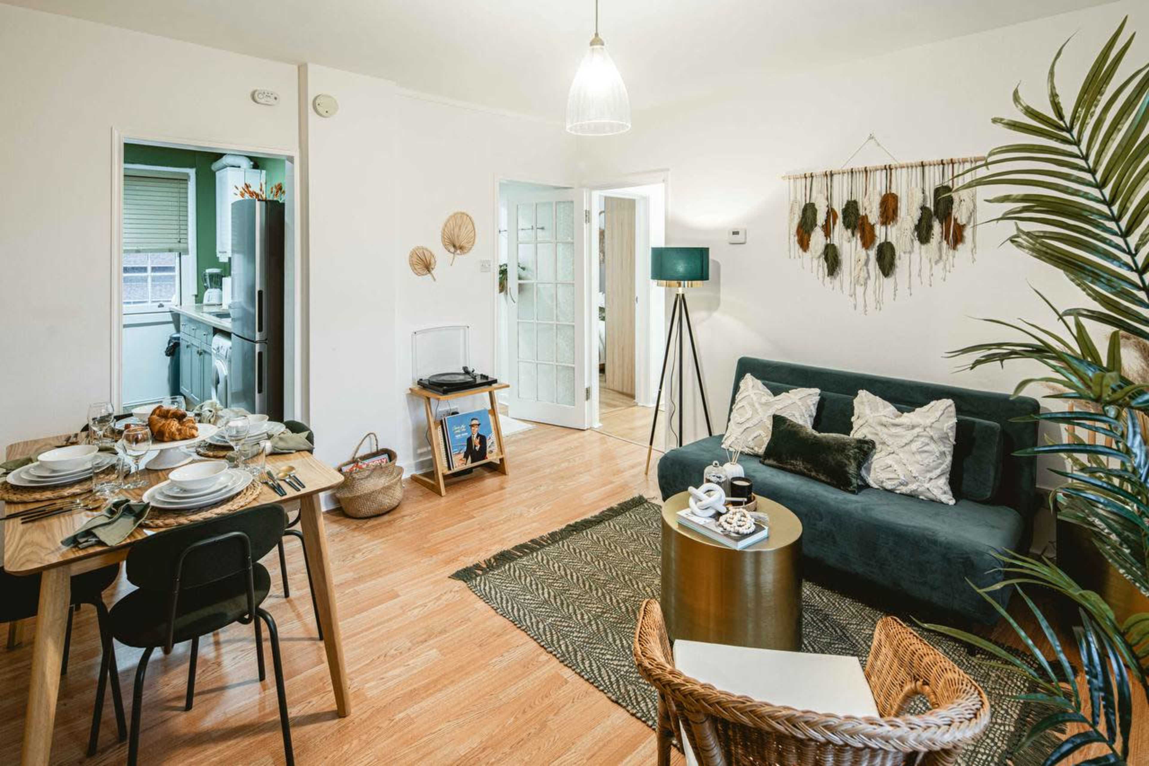 The image shows a cozy living room with a green sofa, a dining table set for a meal, and a decorative wall hanging.