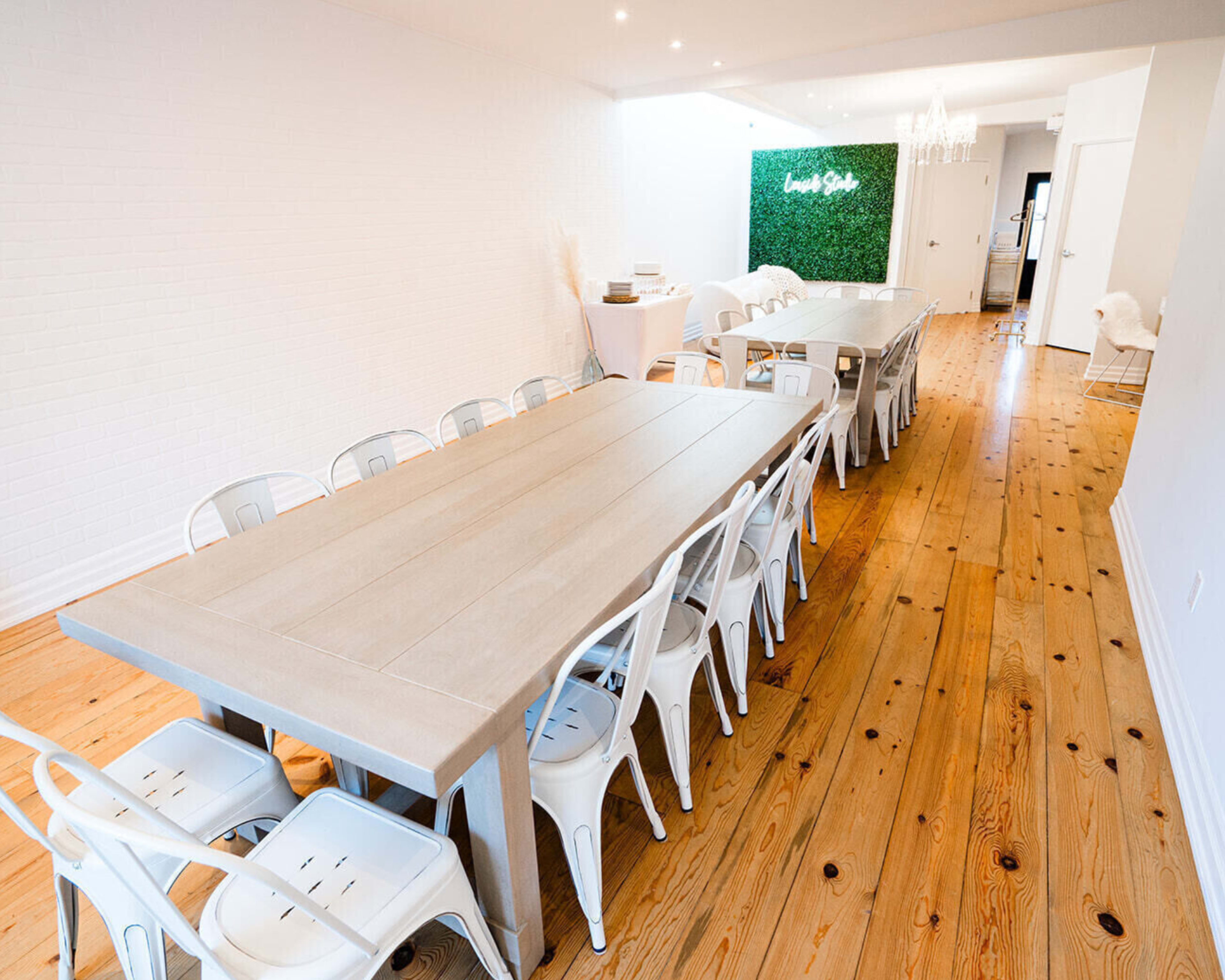 A long, airy dining room features two large wooden tables surrounded by white metal chairs, with a green wall sign in the background.