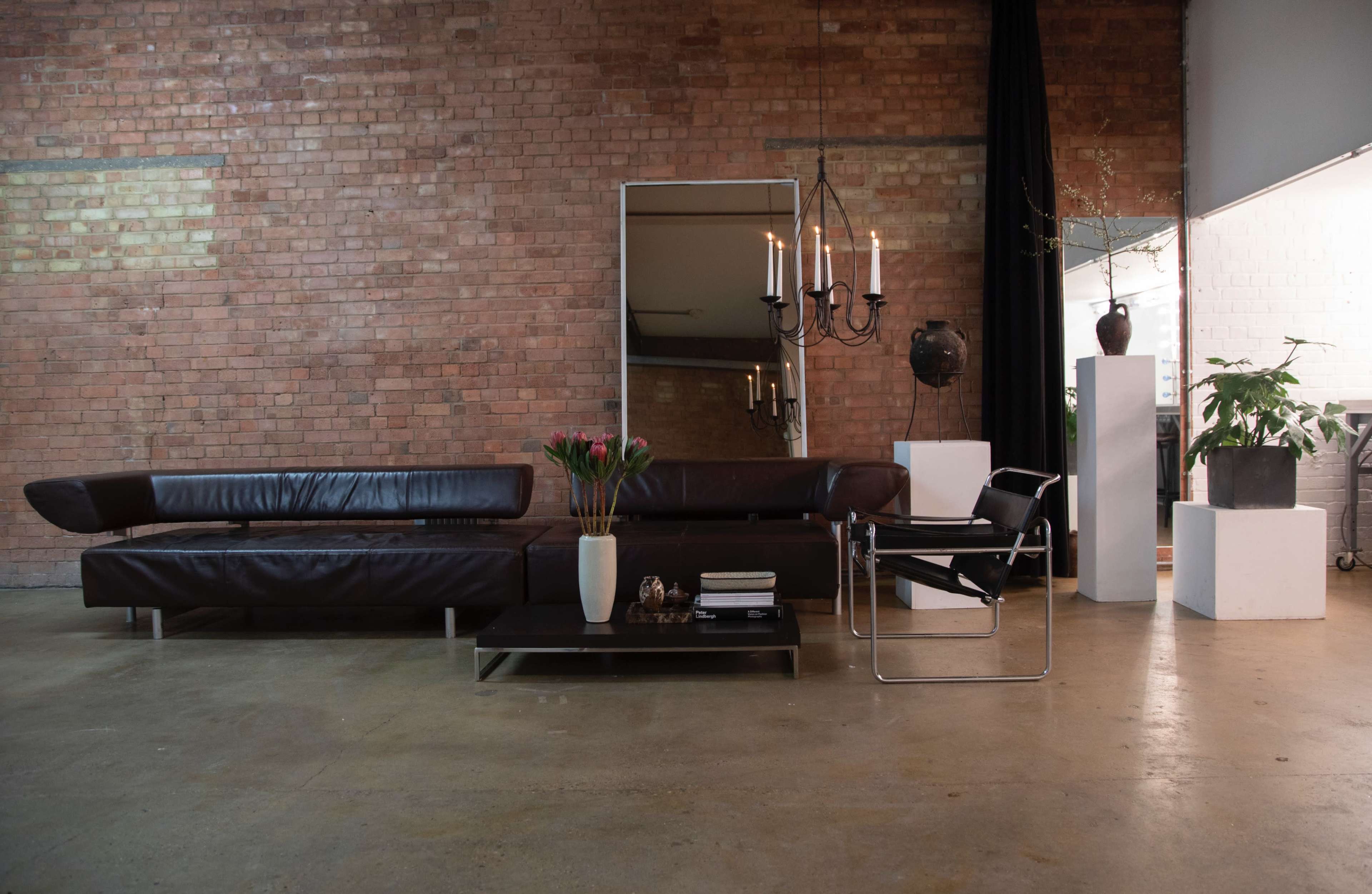 A modern living space featuring a dark leather sofa, a glass coffee table with decorative items, a large mirror, and plants against a brick wall.