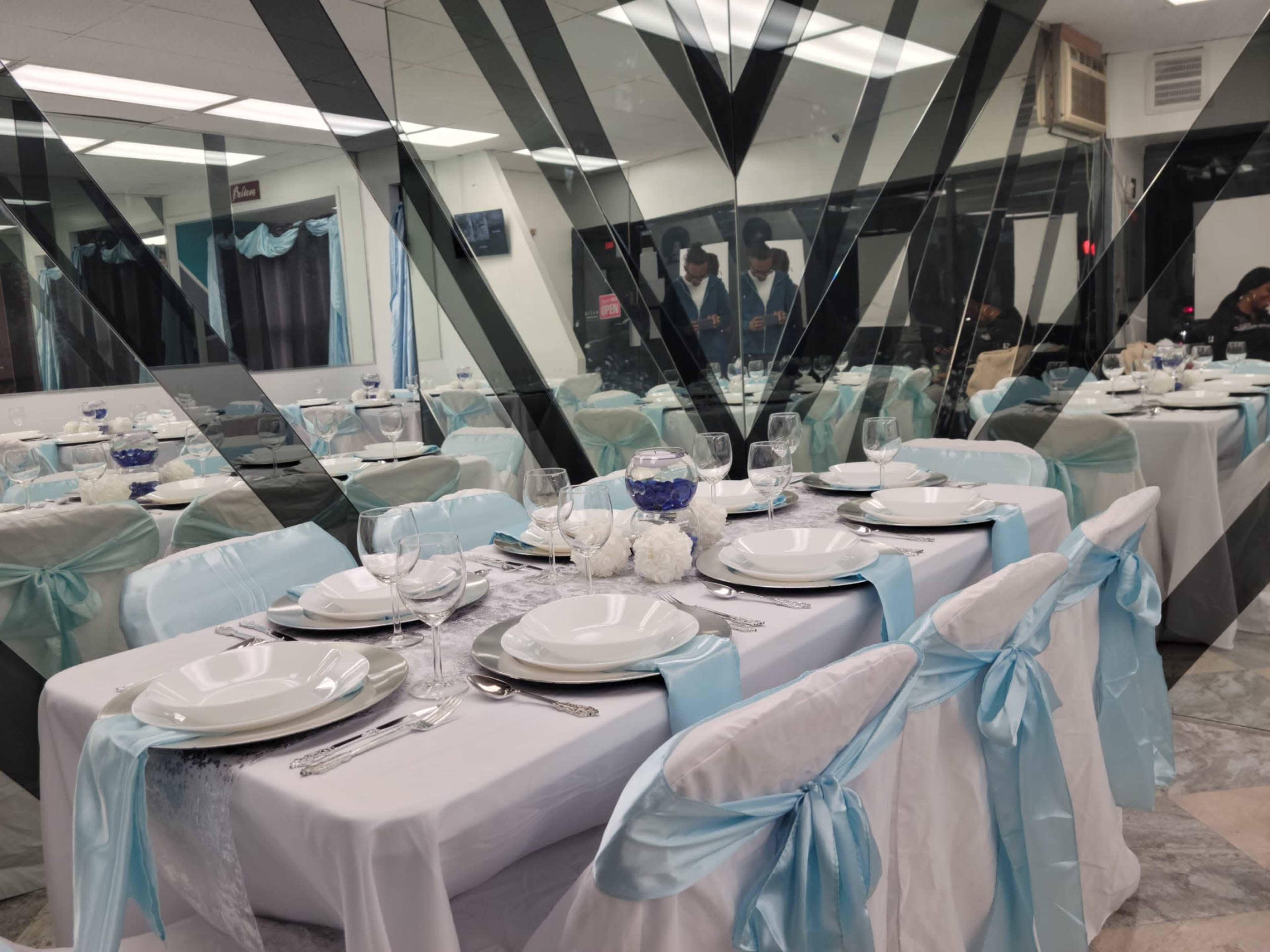 A banquet hall is set up with tables covered in white linens and light blue sashes, featuring centerpieces and place settings in preparation for an event.