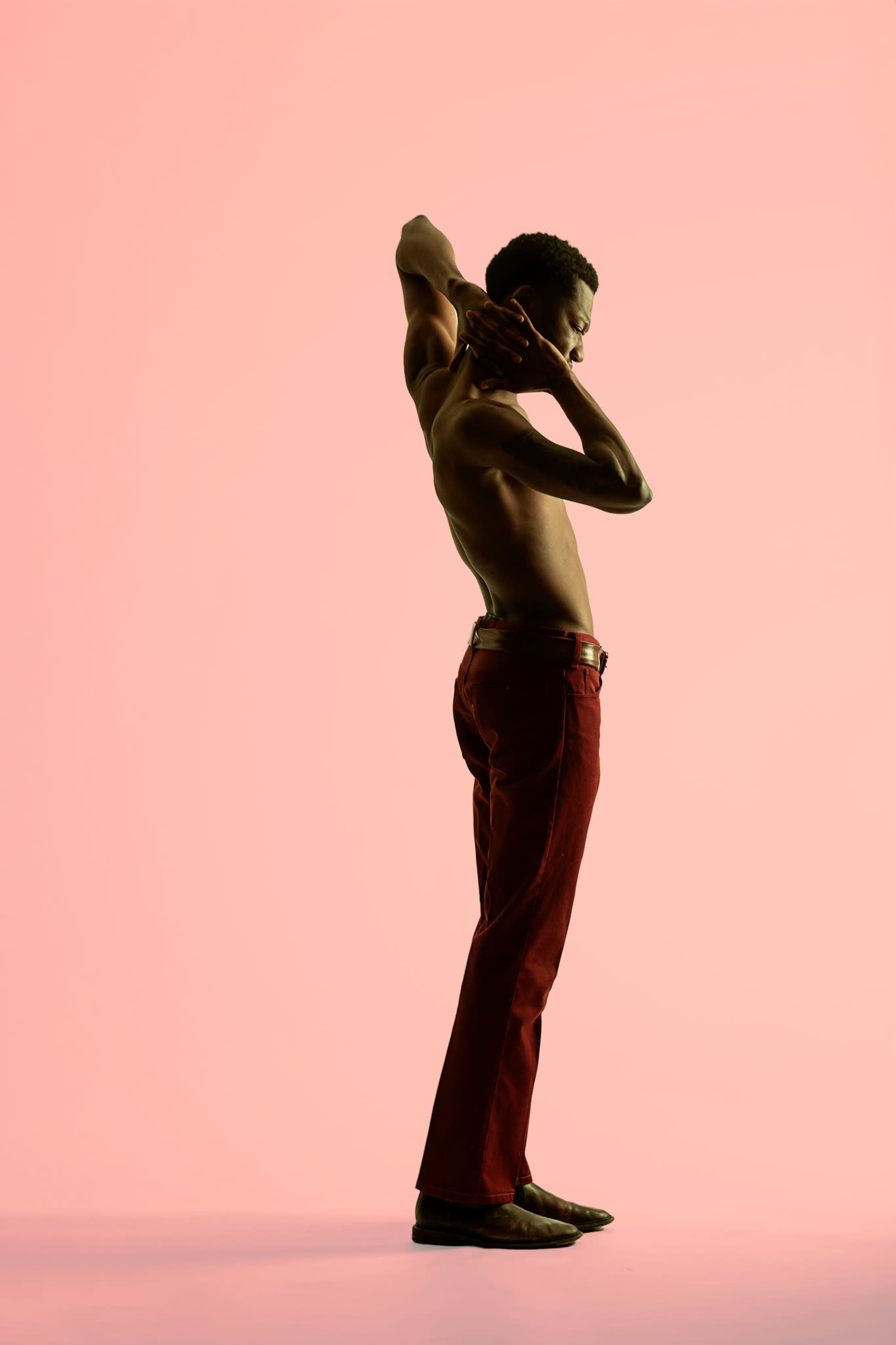 A shirtless man stands in profile against a pink background, stretching his arms behind his head.