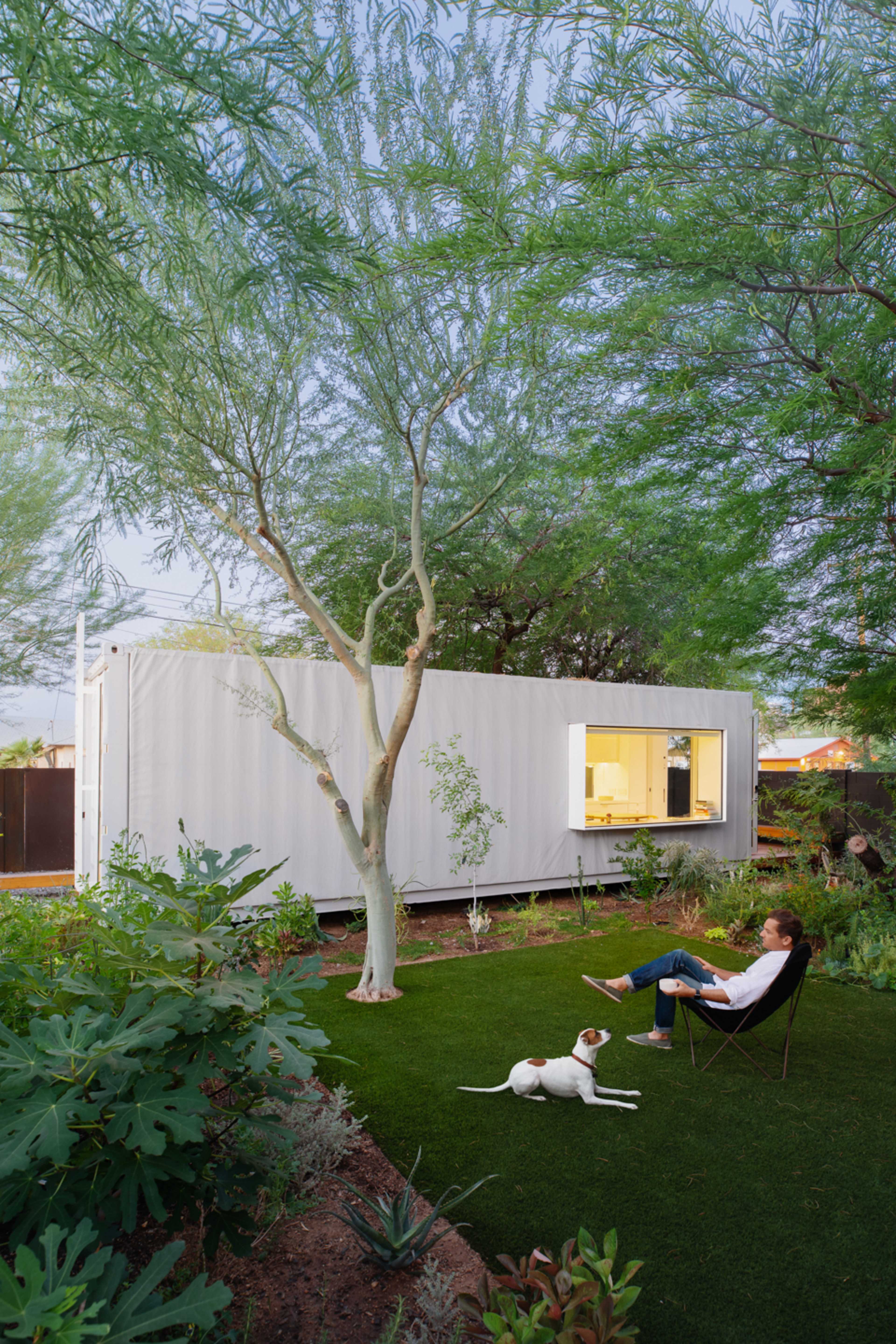 A person sits in a chair next to a white shipping container home surrounded by greenery and a dog lying on the grass.