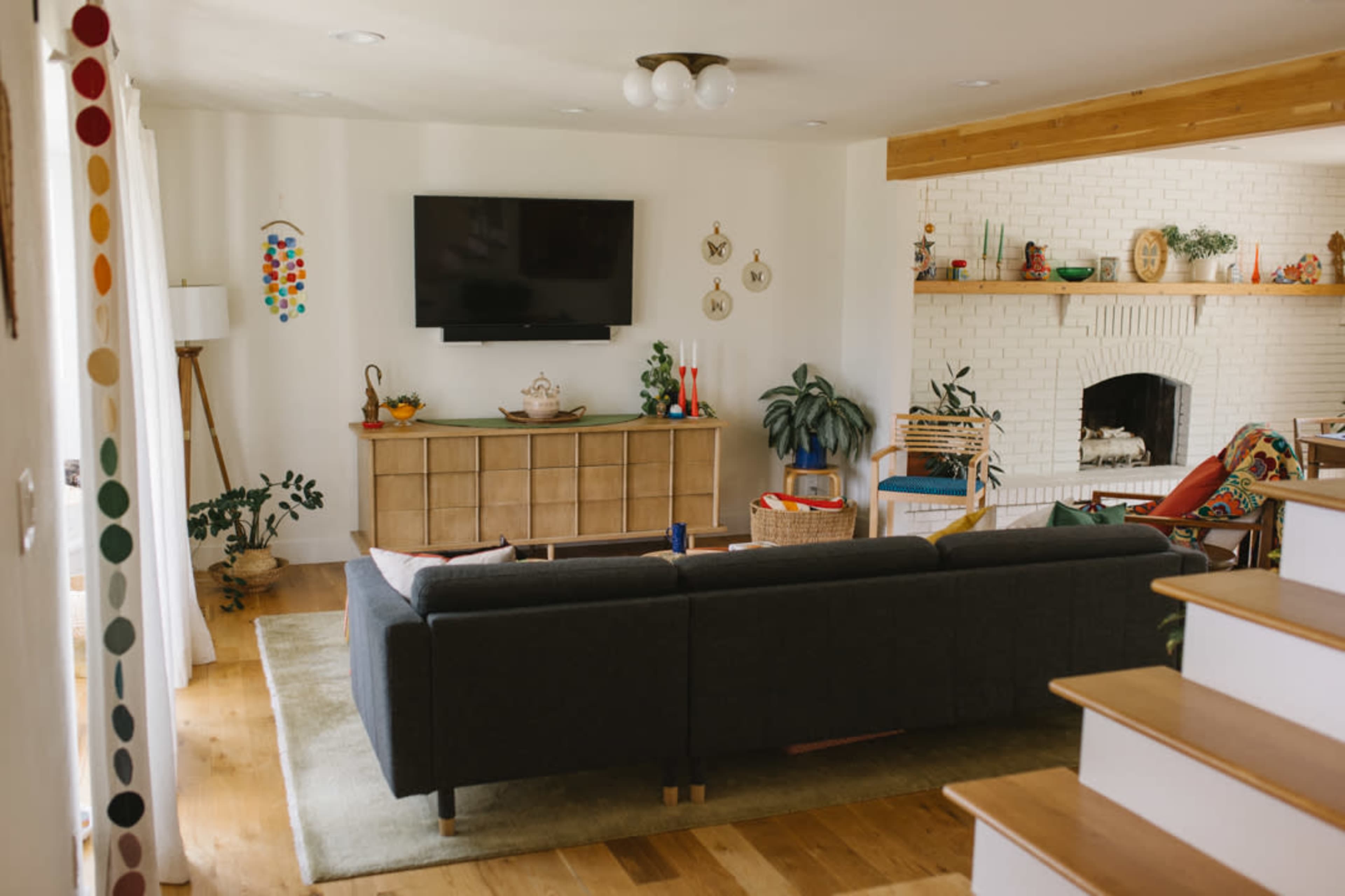 The image shows a modern living room featuring a sectional sofa, a wall-mounted television, a wooden console, and a fireplace in a cozy, well-lit space.