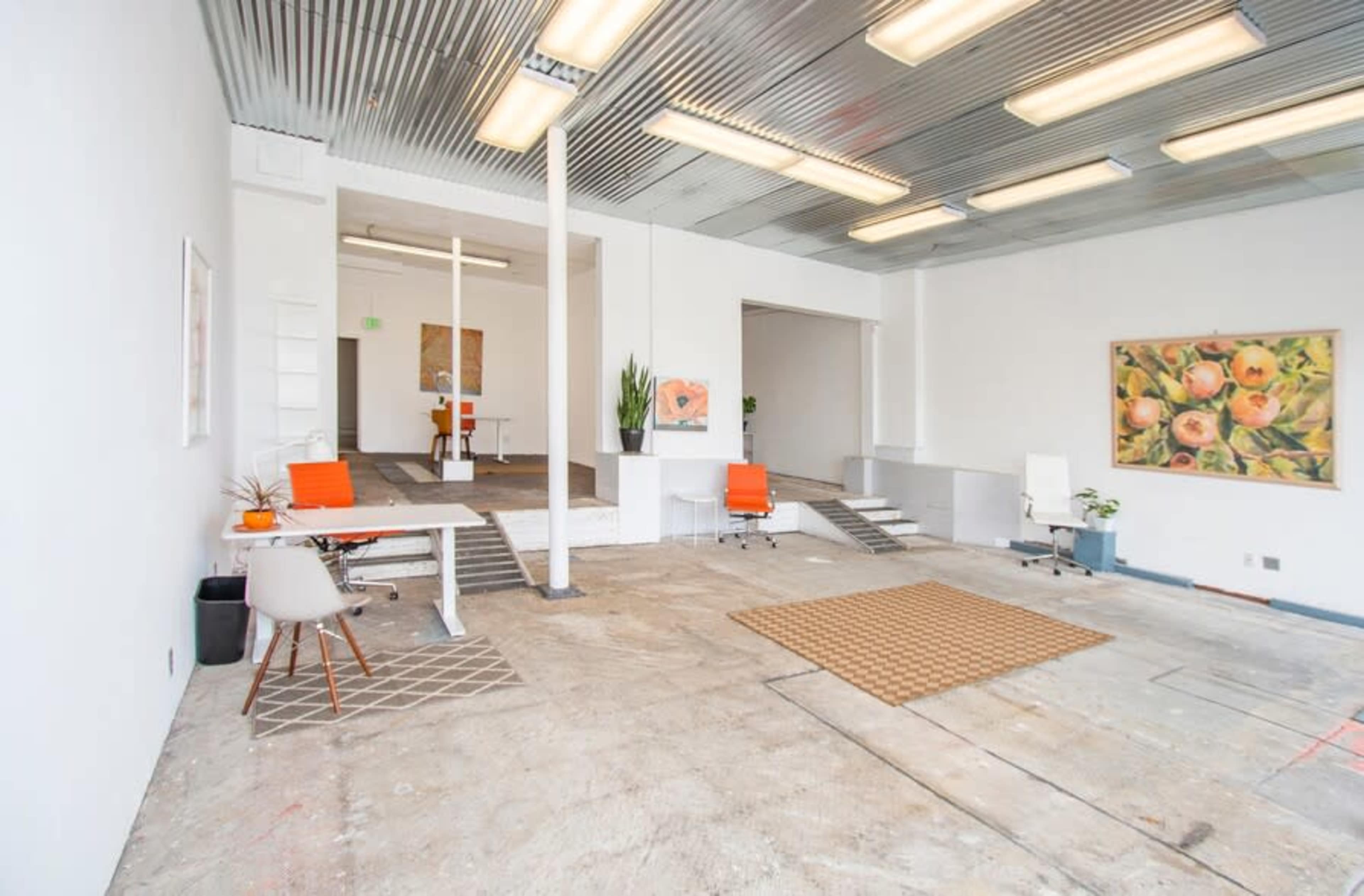 The image shows an open, modern workspace with concrete flooring, orange chairs, and a large artwork on the wall.
