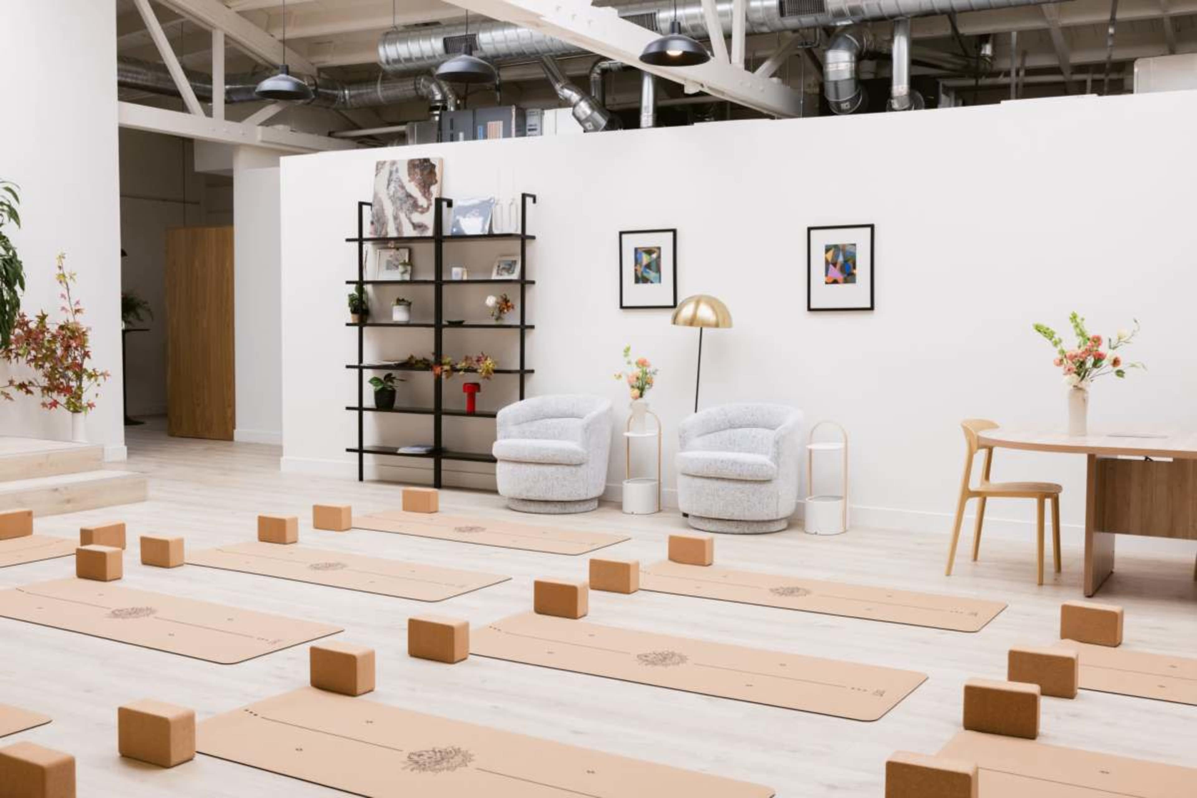 The image shows a minimalist yoga studio with several mats laid out on the floor, wooden blocks placed next to them, and a few decorative elements such as plants and framed pictures on the walls.