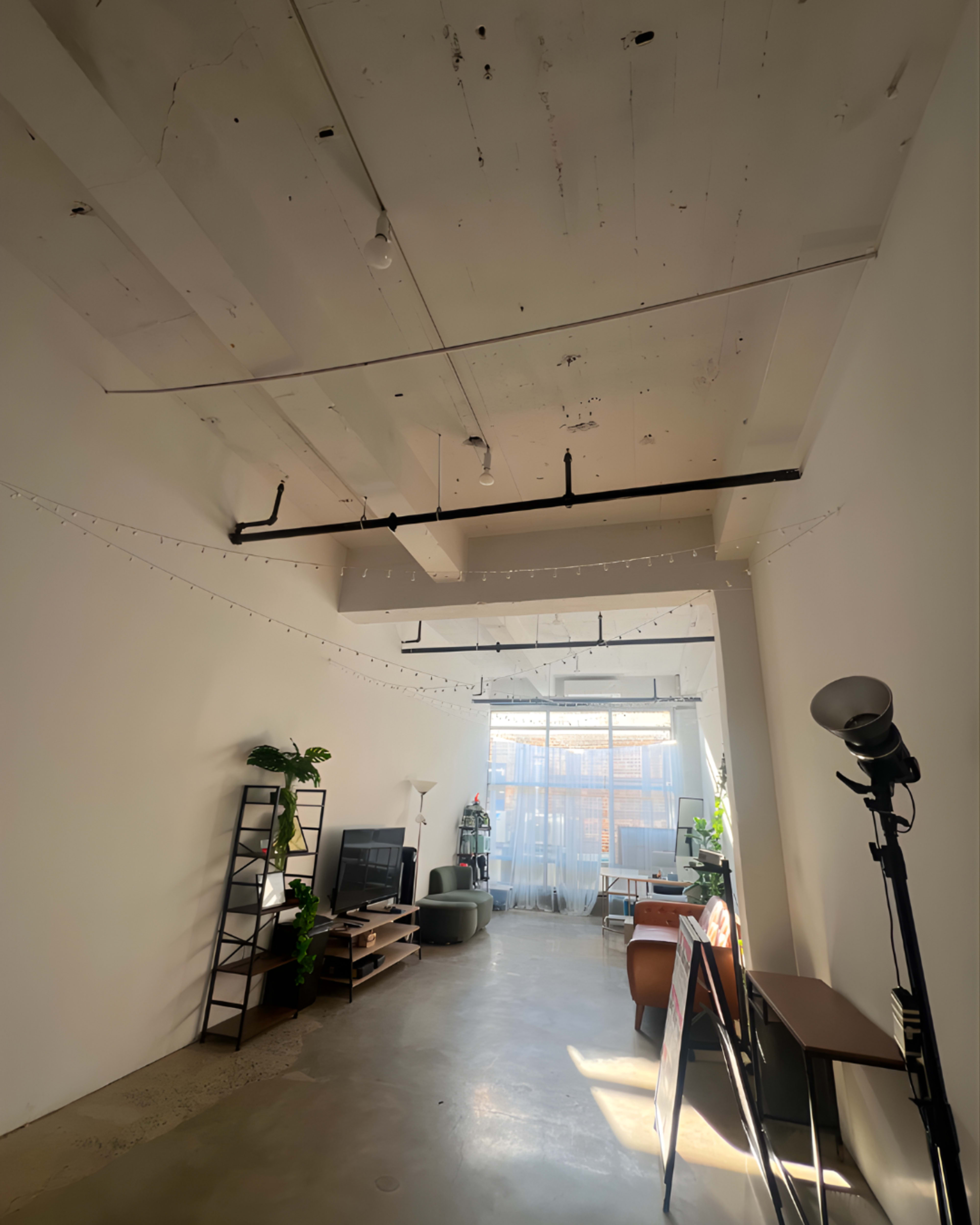 The image shows a spacious room with a concrete floor, large windows allowing natural light, and minimal furnishings including a bookshelf, a television, and a floor lamp.