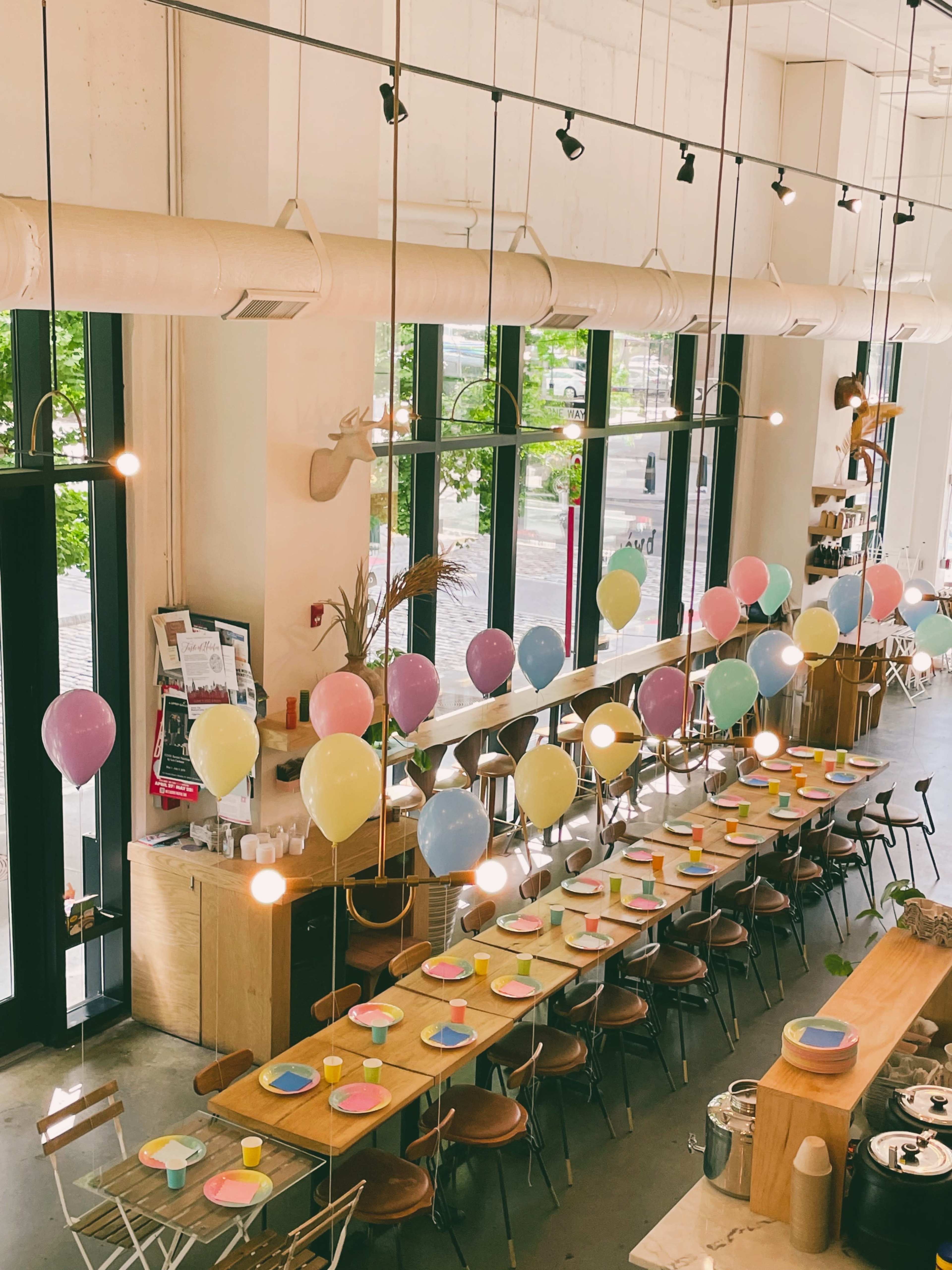 The image shows a spacious café with a long table set for a celebration, featuring pastel-colored plates and an arrangement of balloons hanging above.