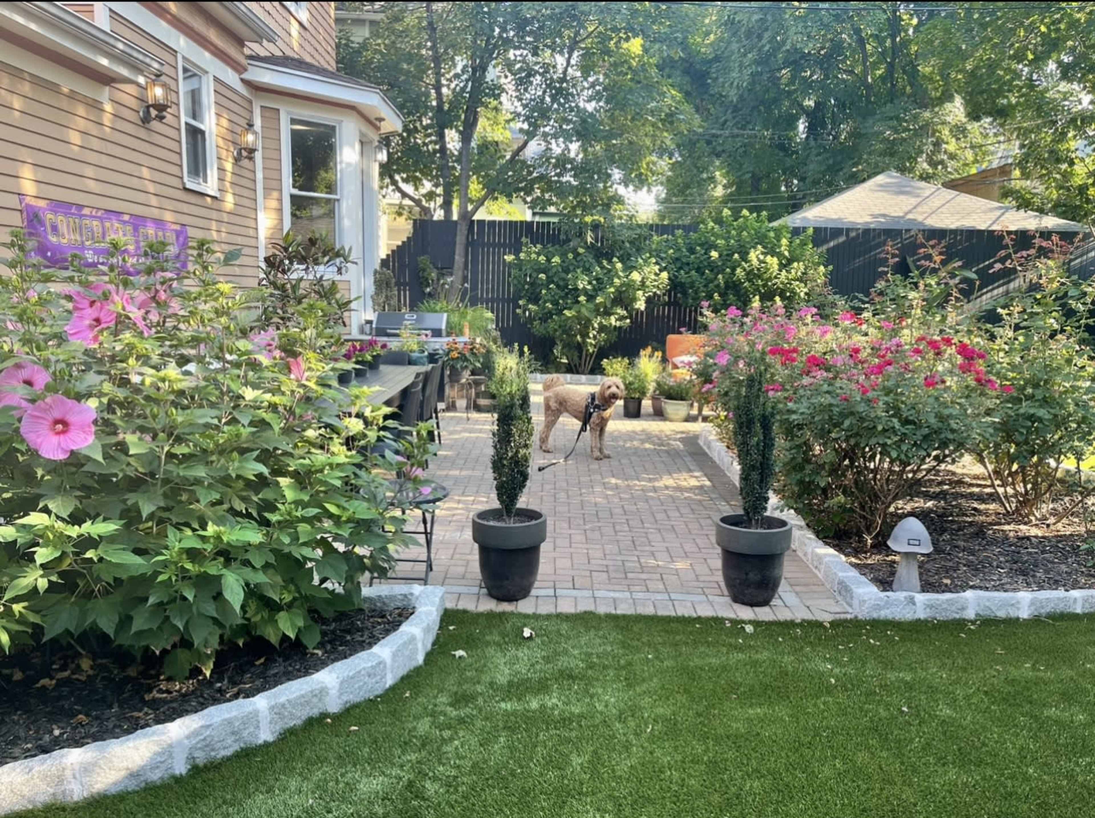 A landscaped backyard features a brick patio surrounded by flower beds and a dog standing on the patio.