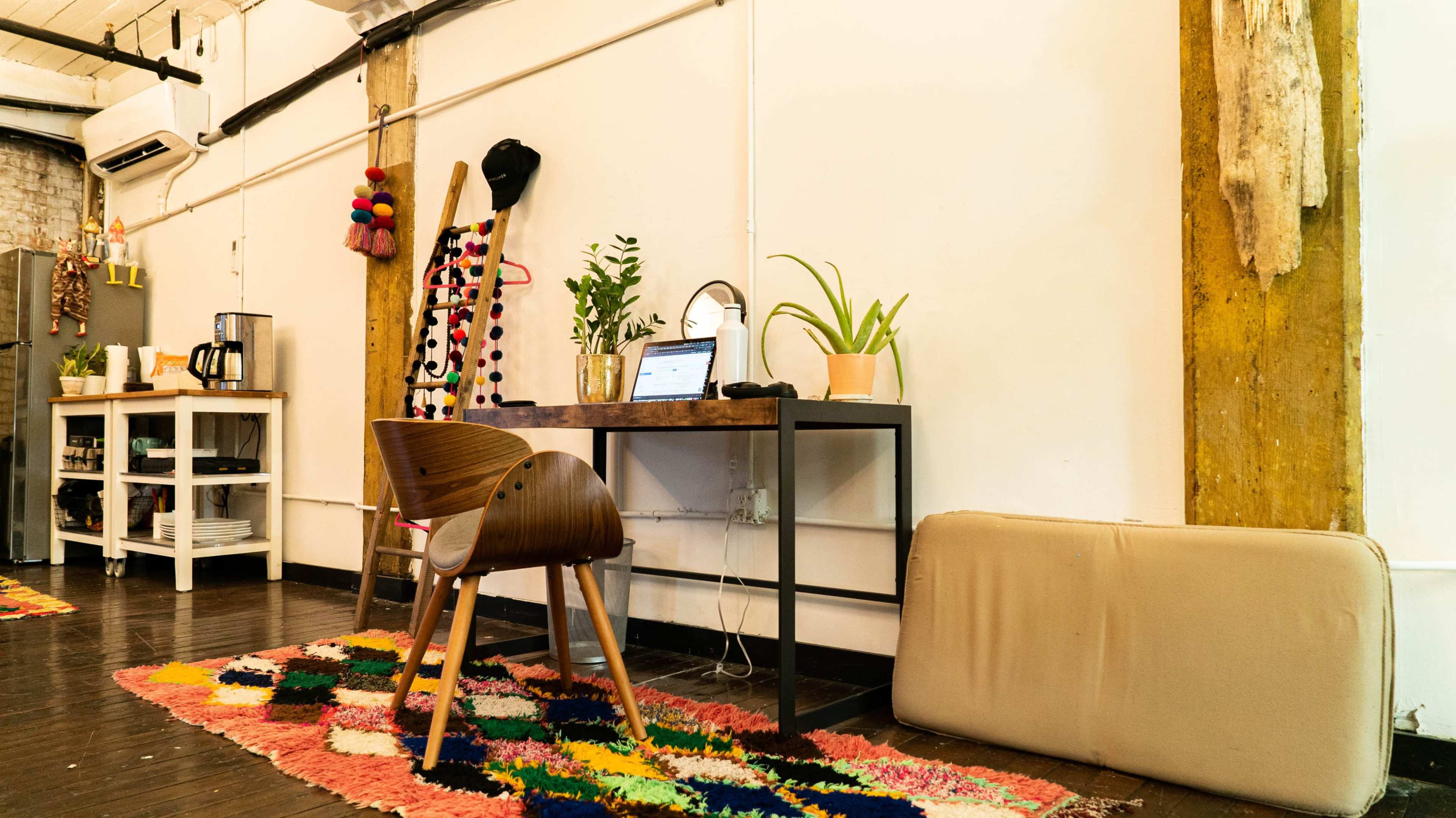 A wooden chair sits at a desk with a laptop, surrounded by a colorful rug and plants in a well-lit room with exposed beams.