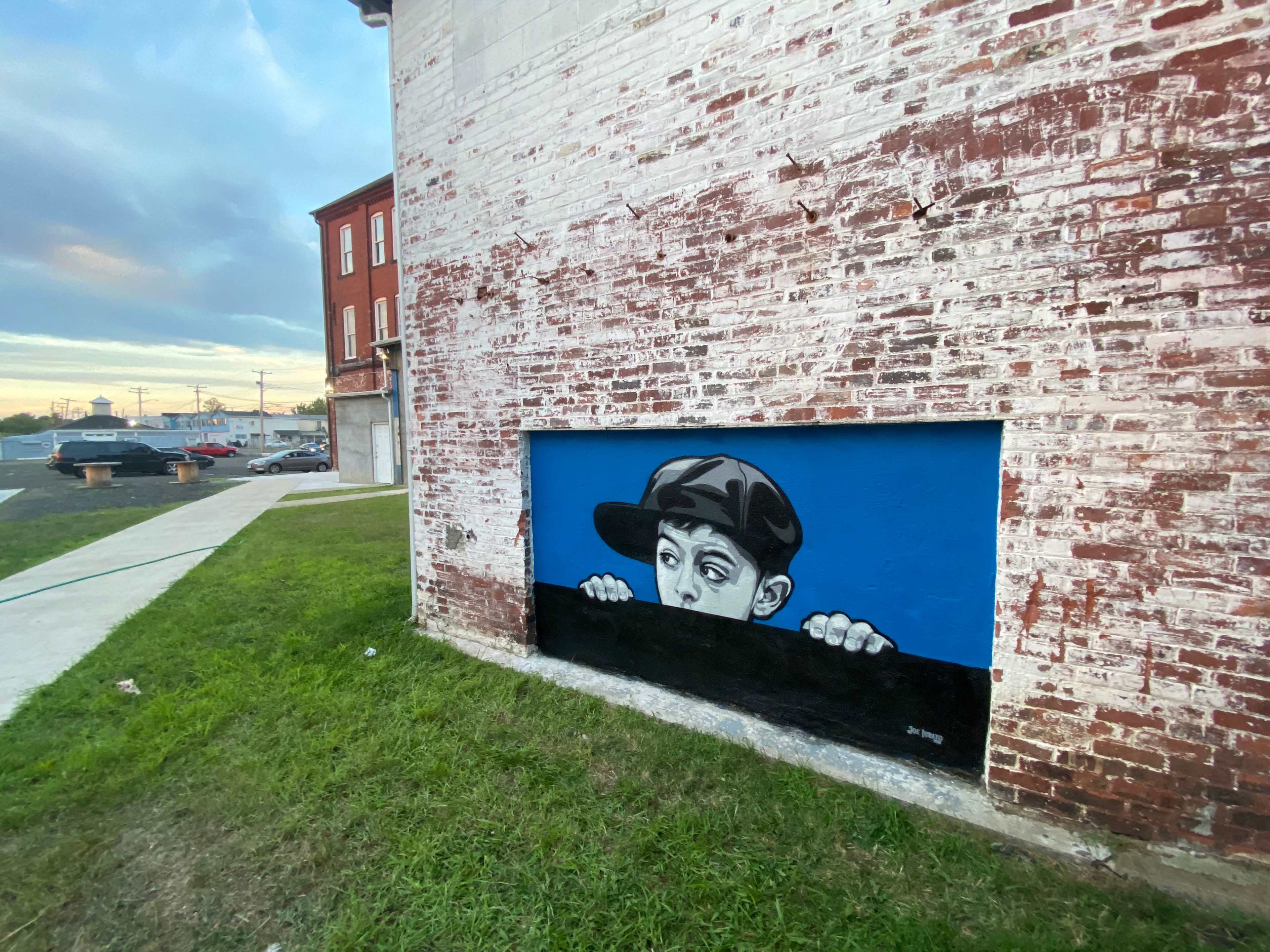A mural depicting a young boy peeking over a wall is painted on a brick building.