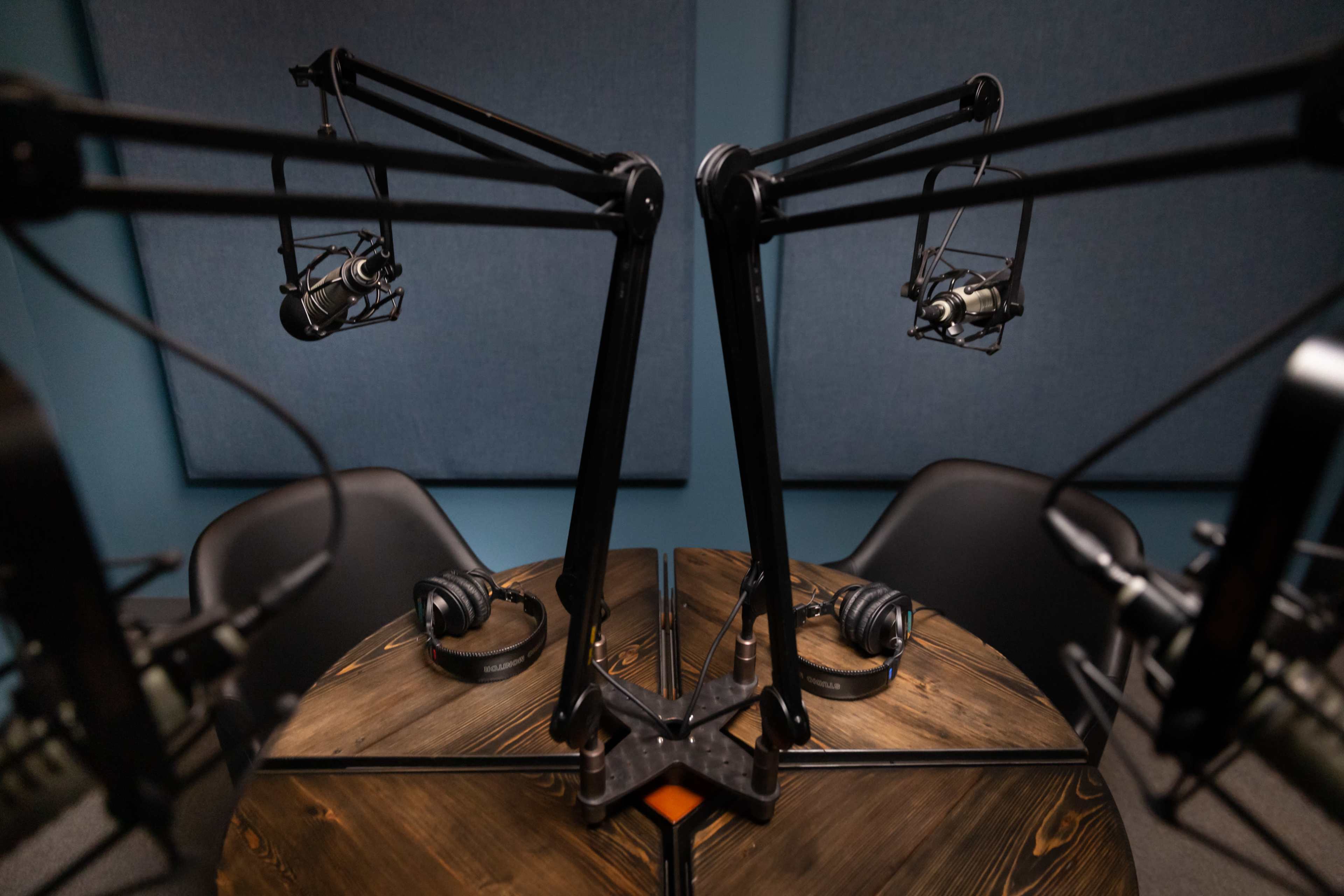 The image shows a podcast recording studio featuring two microphone setups on adjustable stands facing a round wooden table with headphones placed on it.