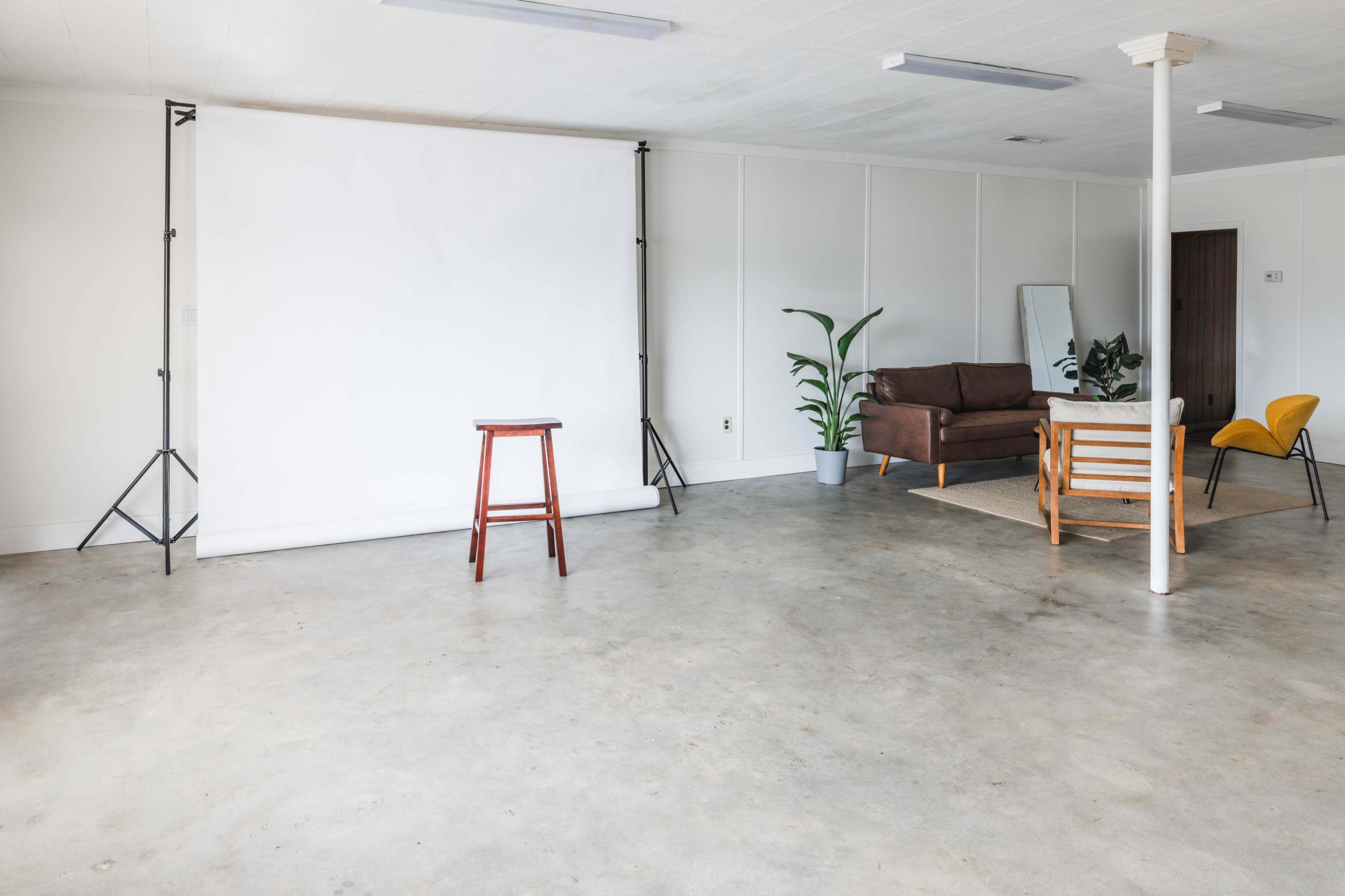 An empty room with a white backdrop, a wooden stool, a brown sofa, a yellow chair, and a potted plant.