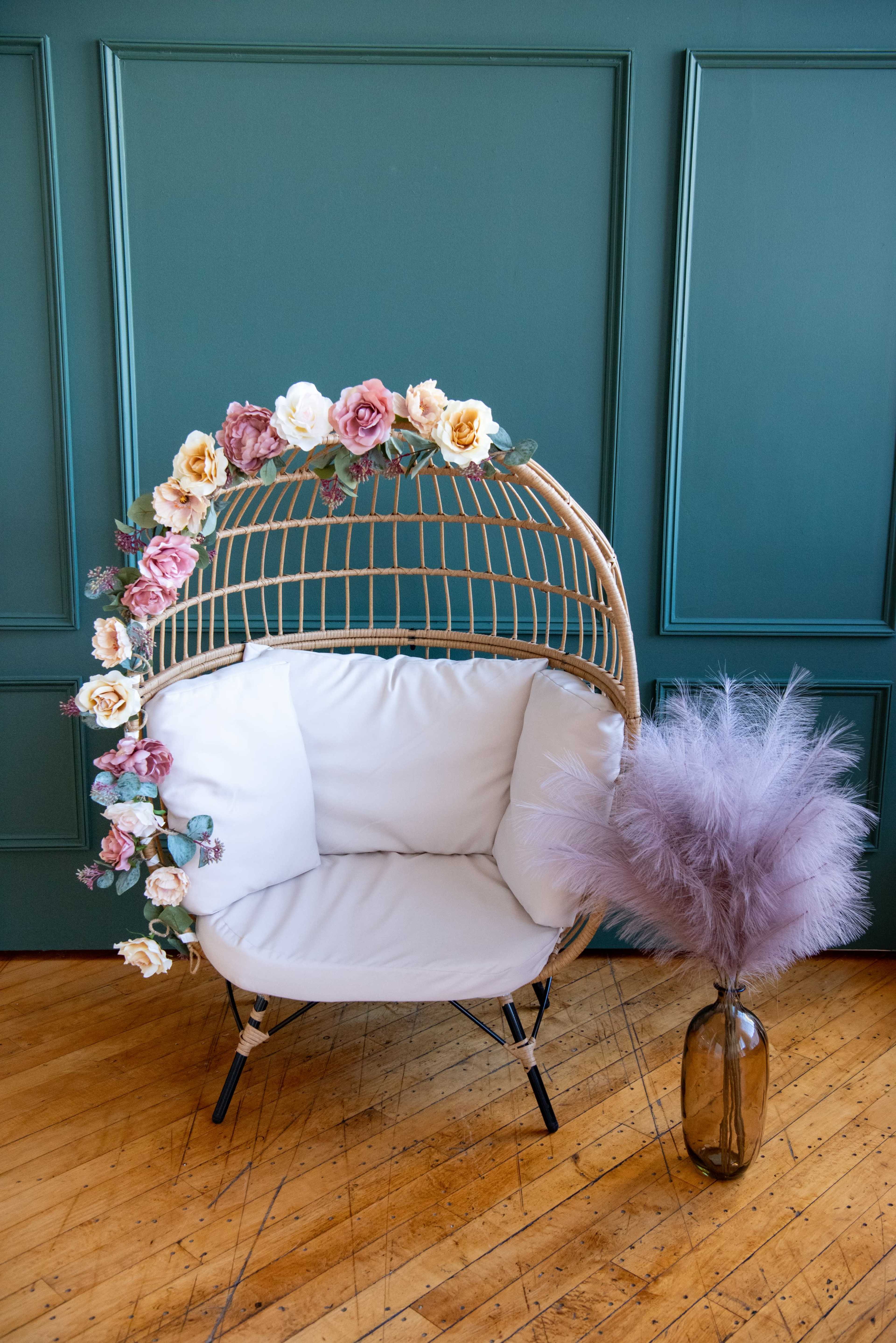 A rattan chair adorned with artificial flowers sits next to a glass vase containing fluffy purple pampas grass against a green wall.
