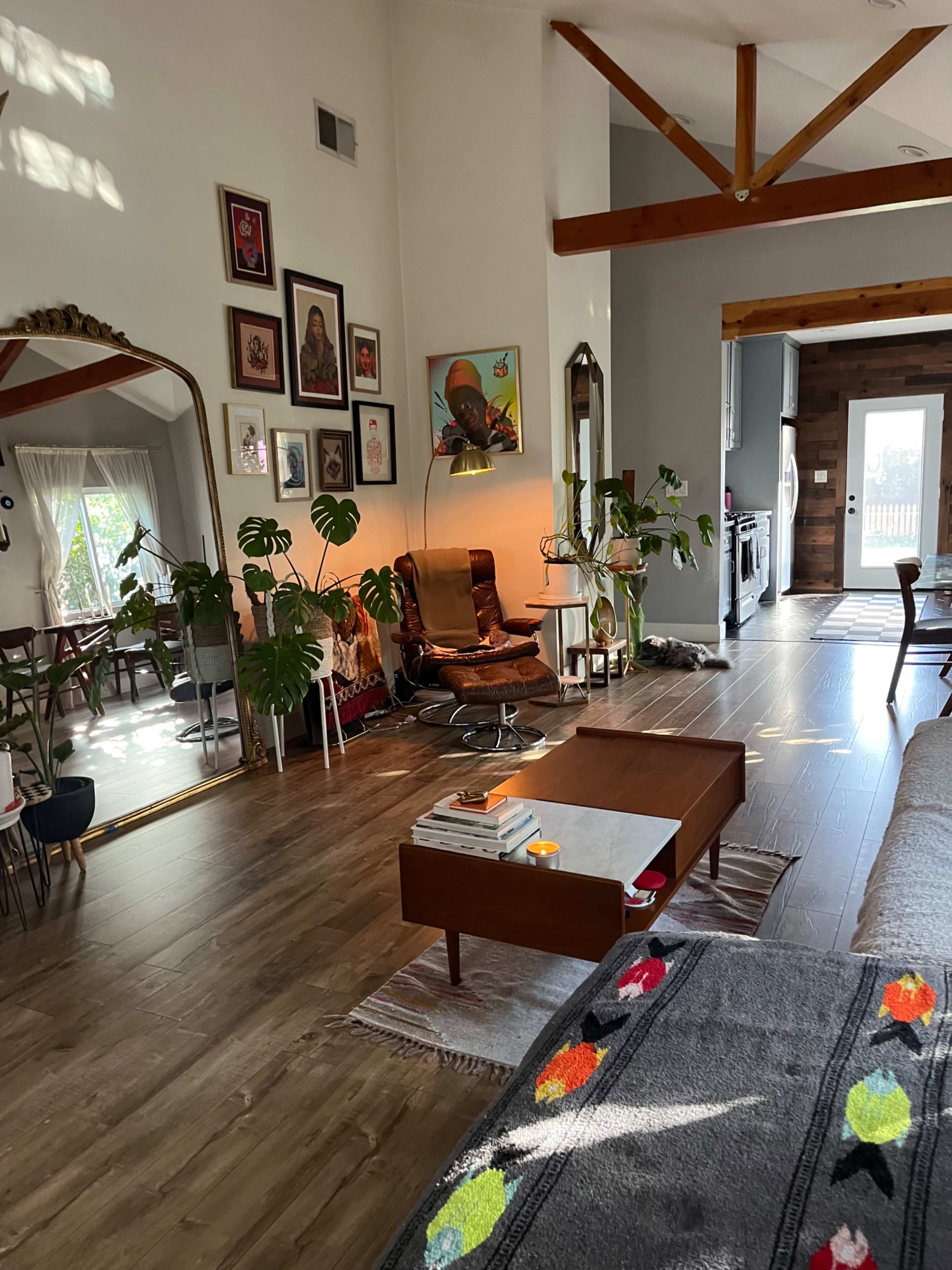 The image shows a spacious, well-lit living area featuring a mix of plants, artwork on the walls, a mid-century modern chair, and a coffee table with decorative items.