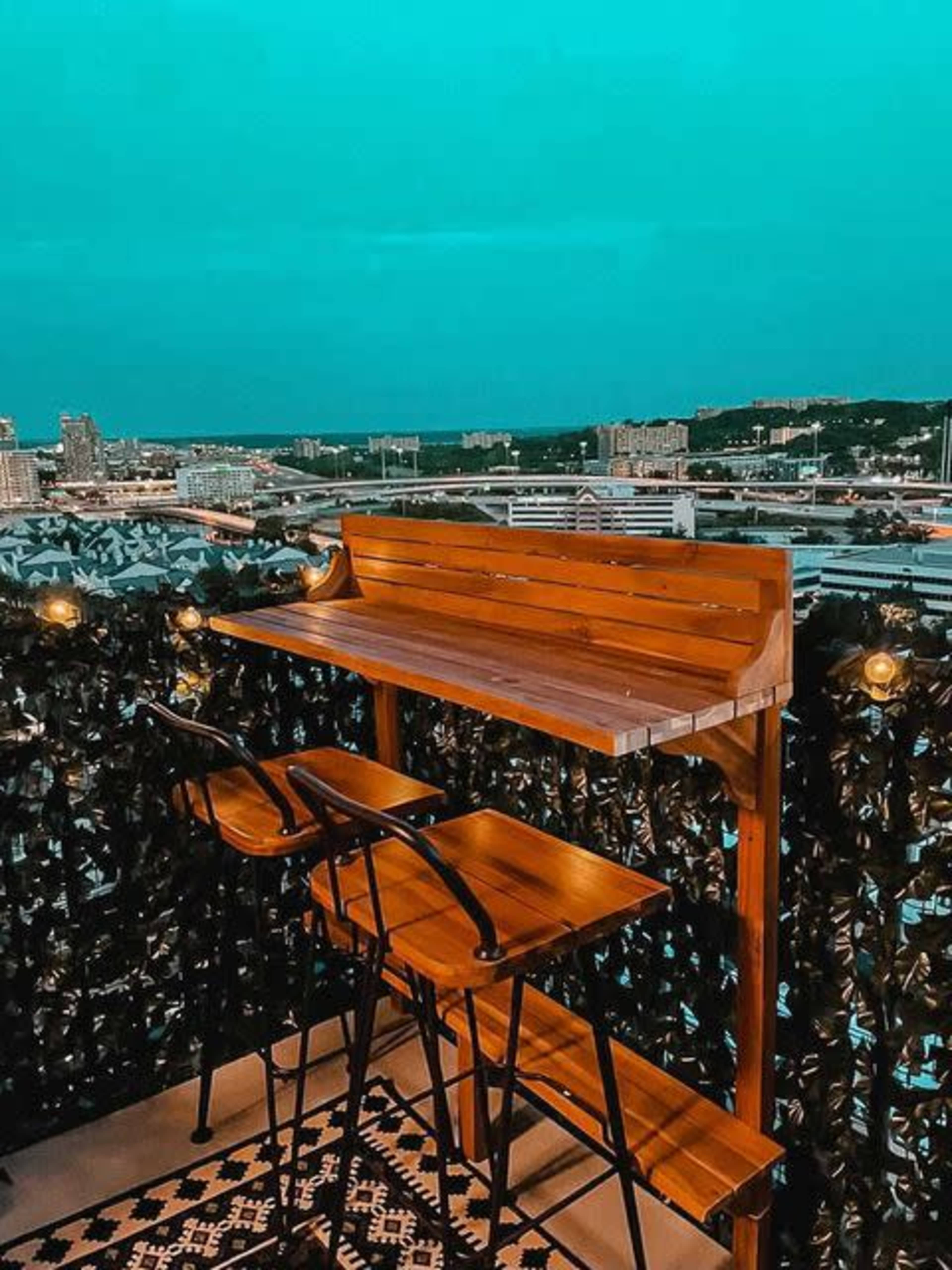 A wooden bar table with three stools overlooks a city skyline under a blue sky.