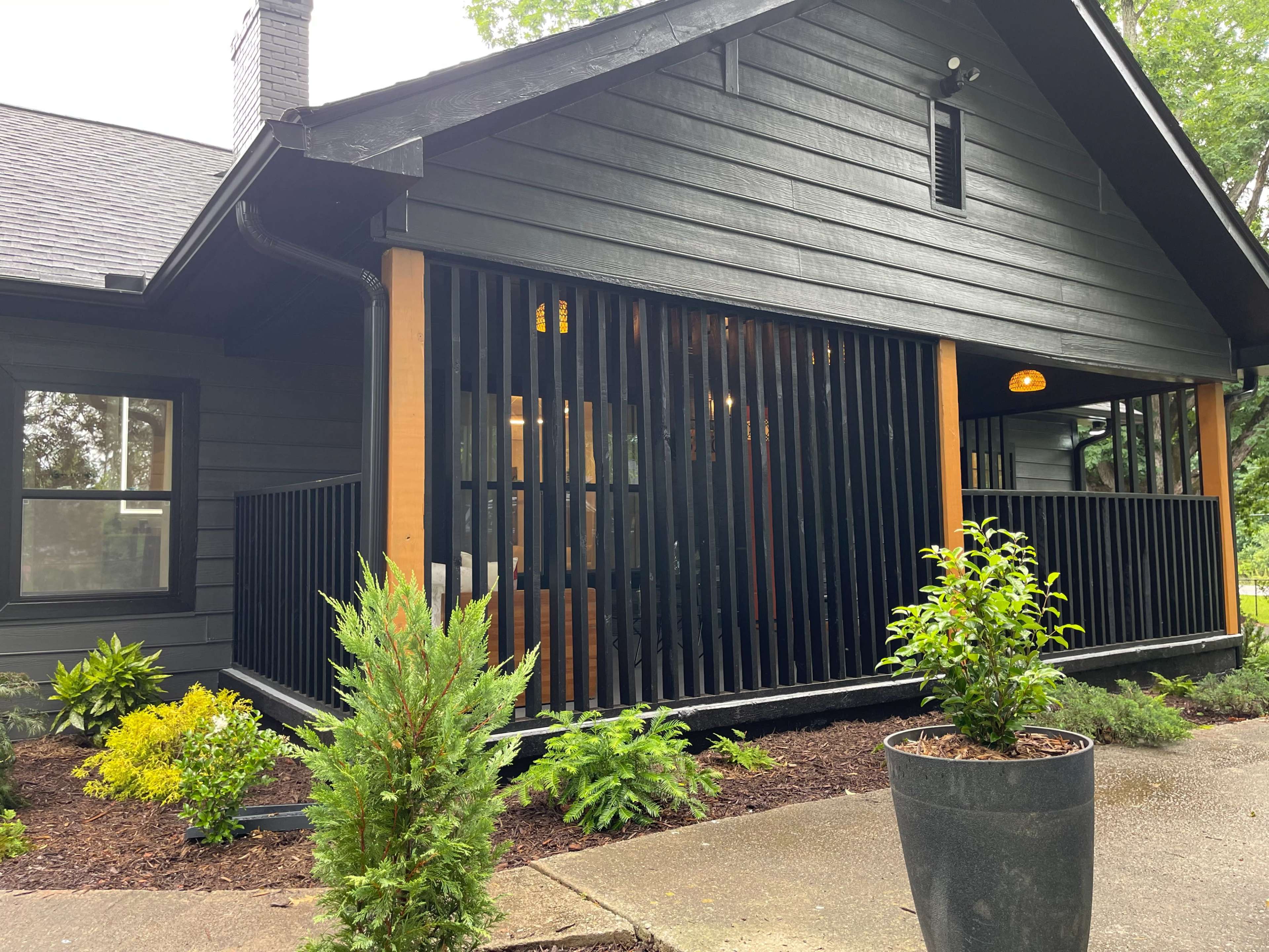 The image shows a modern black house with wooden accents, featuring vertical slatted fencing and landscaped greenery in front.